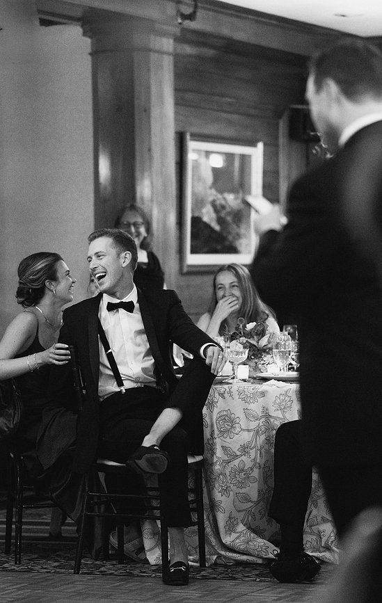 Black and white photo of wedding guests laughing joyfully during a speech at an elegant reception.