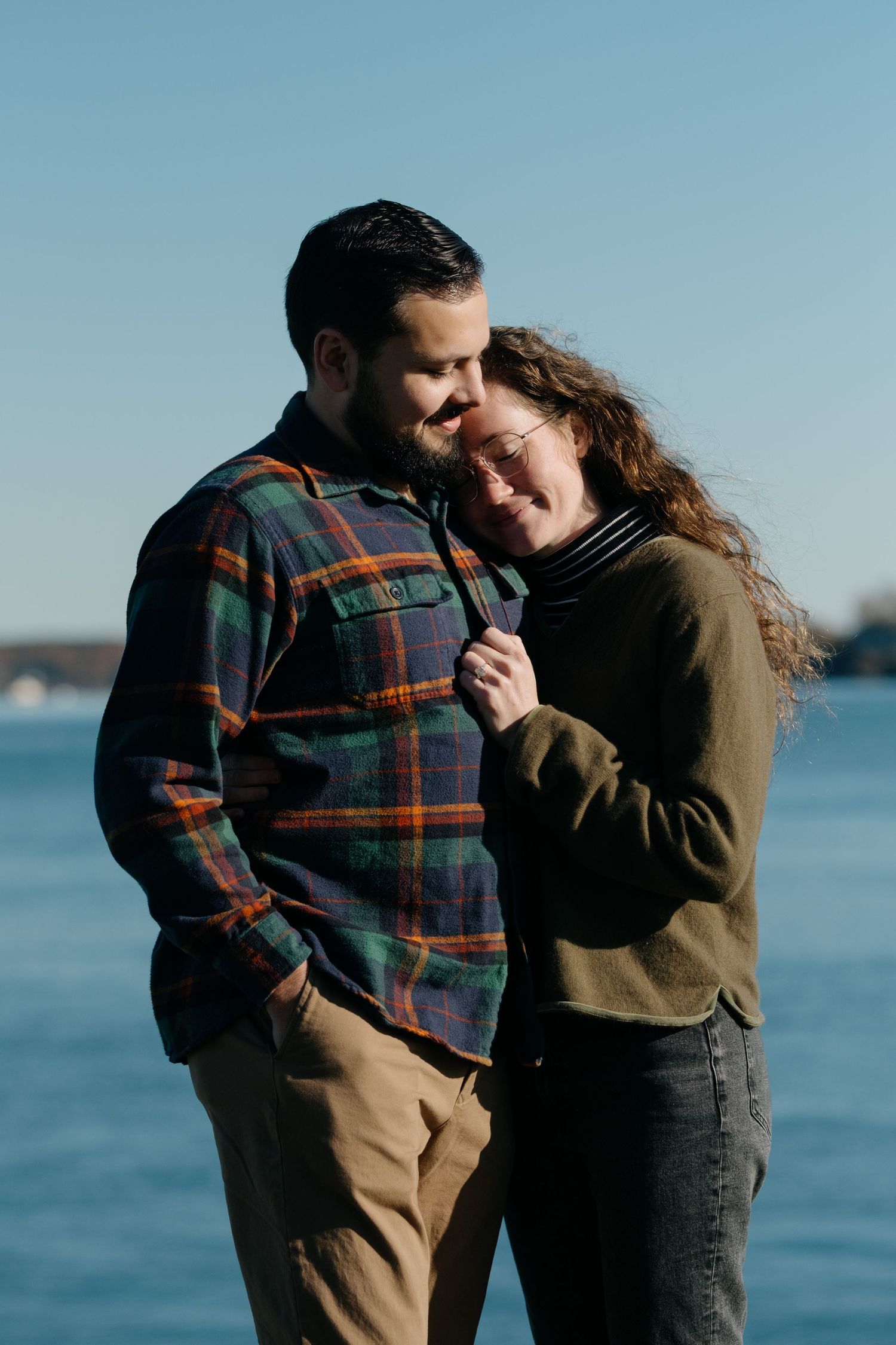 Couple smiles and leans together affectionately with blue water in the background.