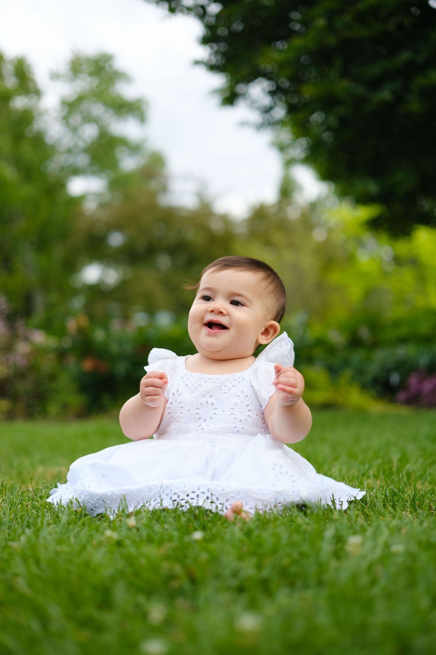 Adorable baby girl in a white dress sitting on green grass smiling joyfully outdoors.