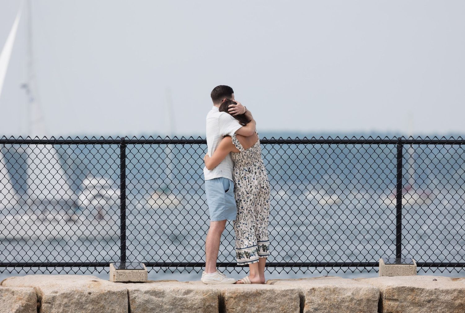 Couple embraces on rocky waterfront with sailboats and harbor visible in background.