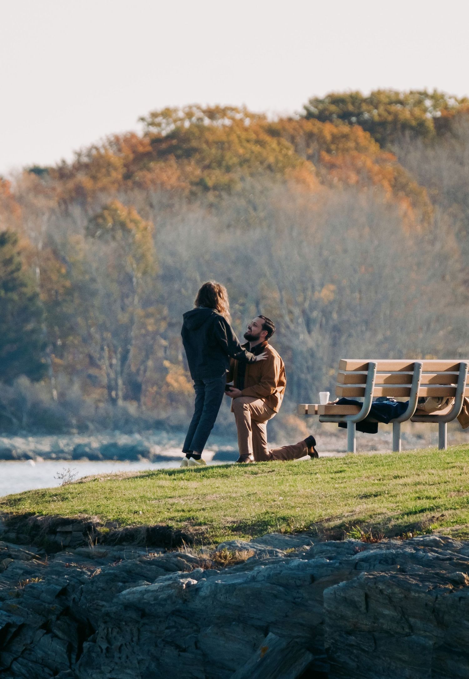 Man proposes on one knee outdoors near rocky shore with autumn foliage behind.