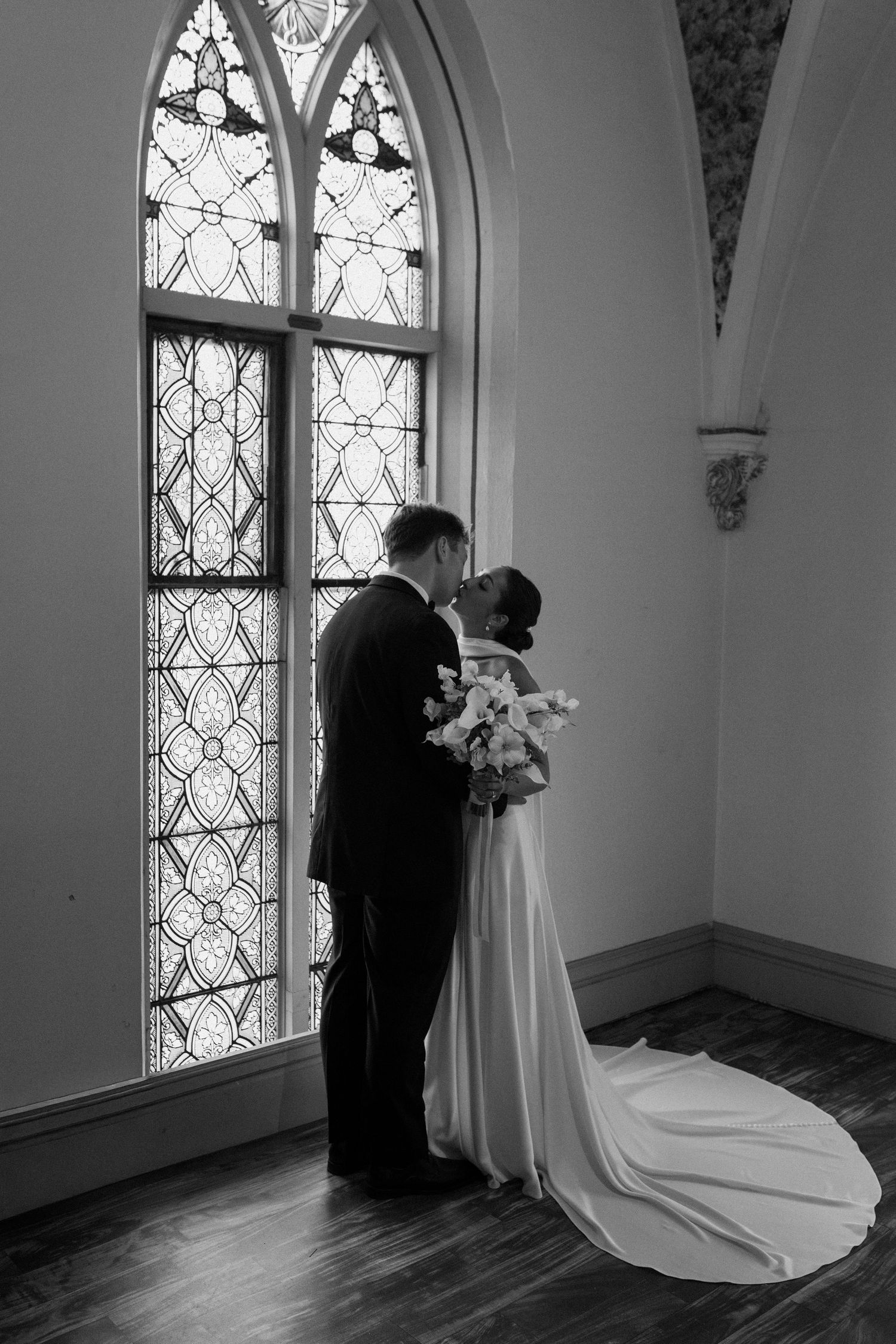 Black and white photo of bride and groom sharing a kiss before a ornate stained glass church window.