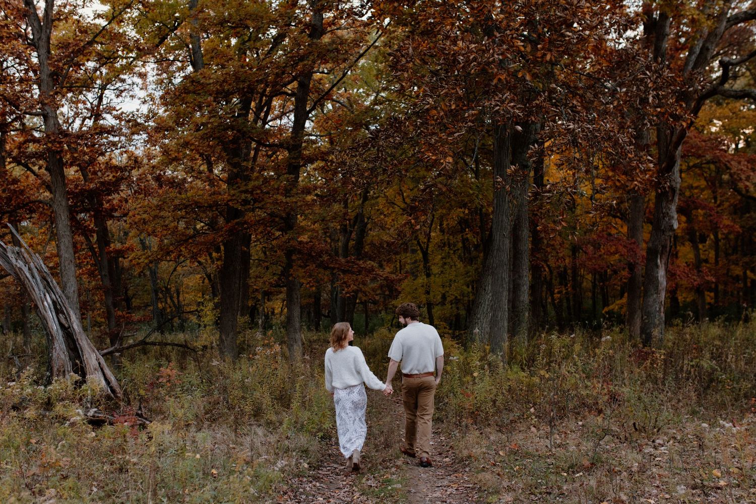 Couple walking hand-in-hand in front of woods at Lapham Peak State Part engagement photography session