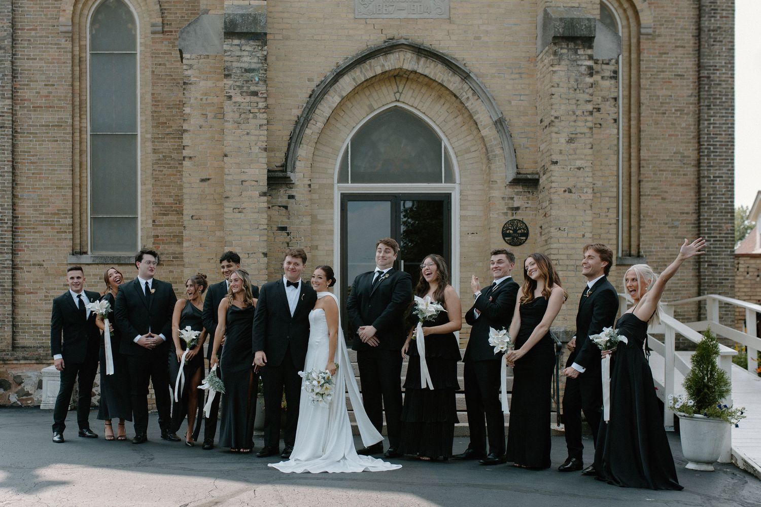 Wedding party in formal black attire poses on church steps for group photo at Gardens Wedding Center in Wisconsin