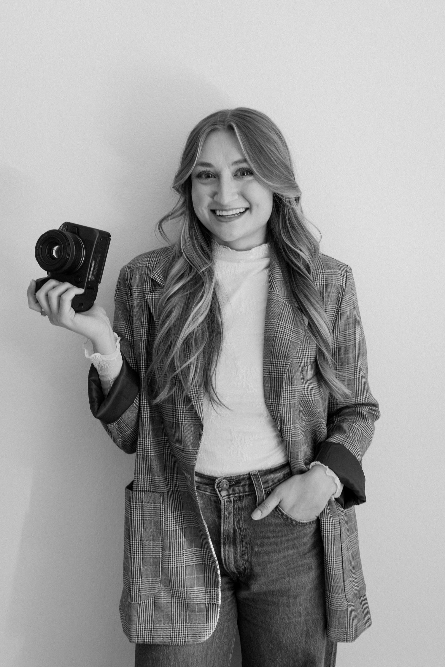Autumn Johnson Photography wears a plaid blazer and white turtleneck poses with a camera in a minimalist studio setting.