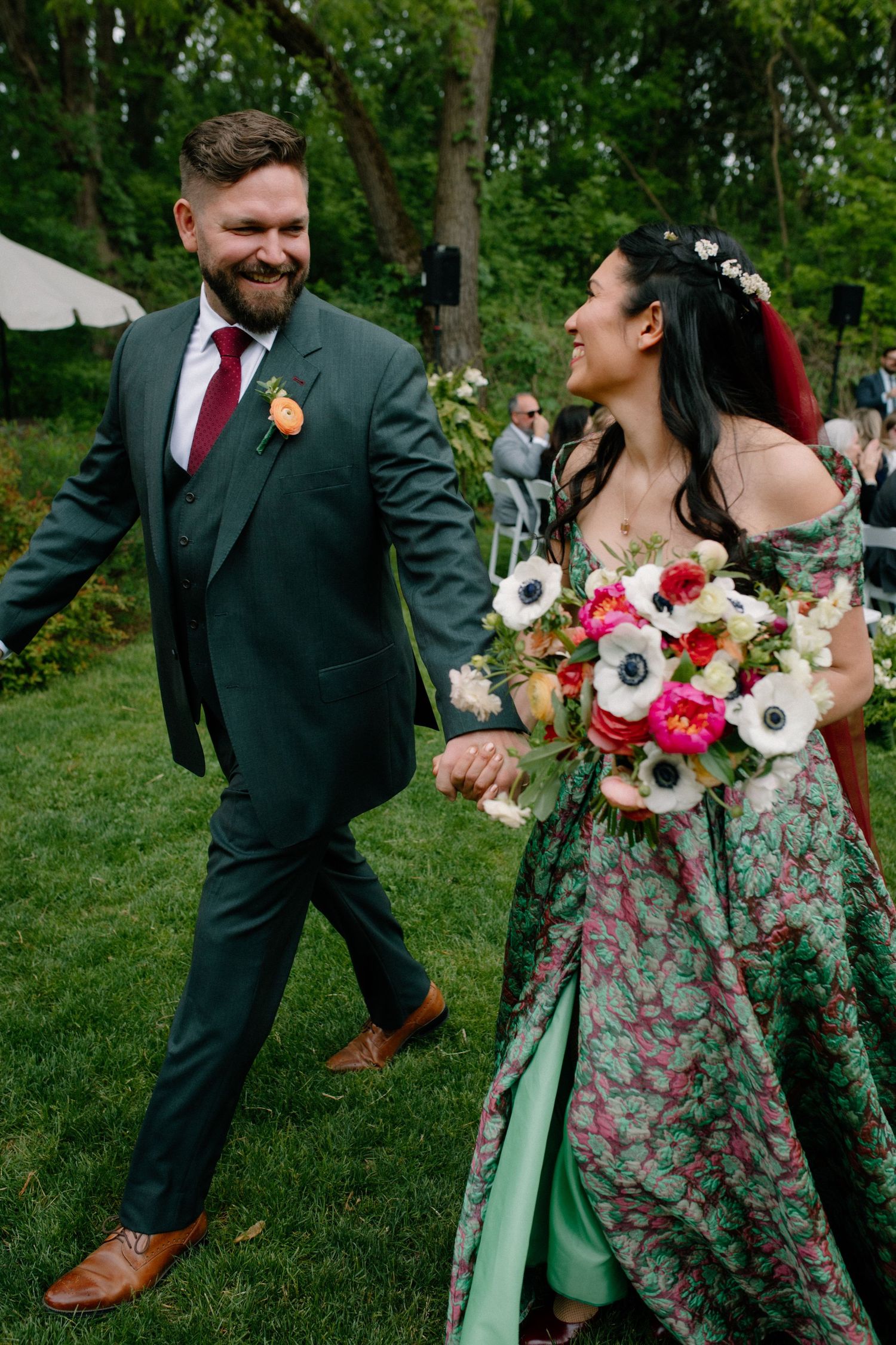 Bride in green floral gown holds bouquet of anemones while walking hand-in-hand with groom in dark green suit.