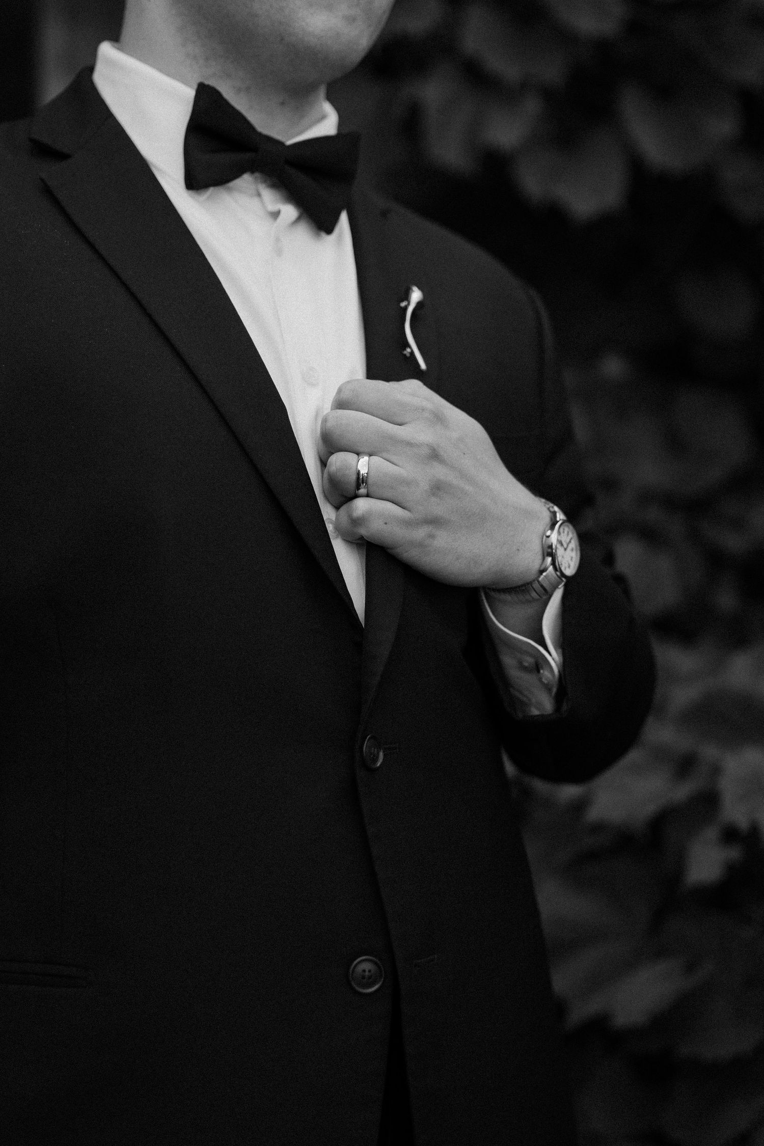 Black and white photo of a groom in a tuxedo with bow tie, adjusting lapel pin, wearing a watch and ring.