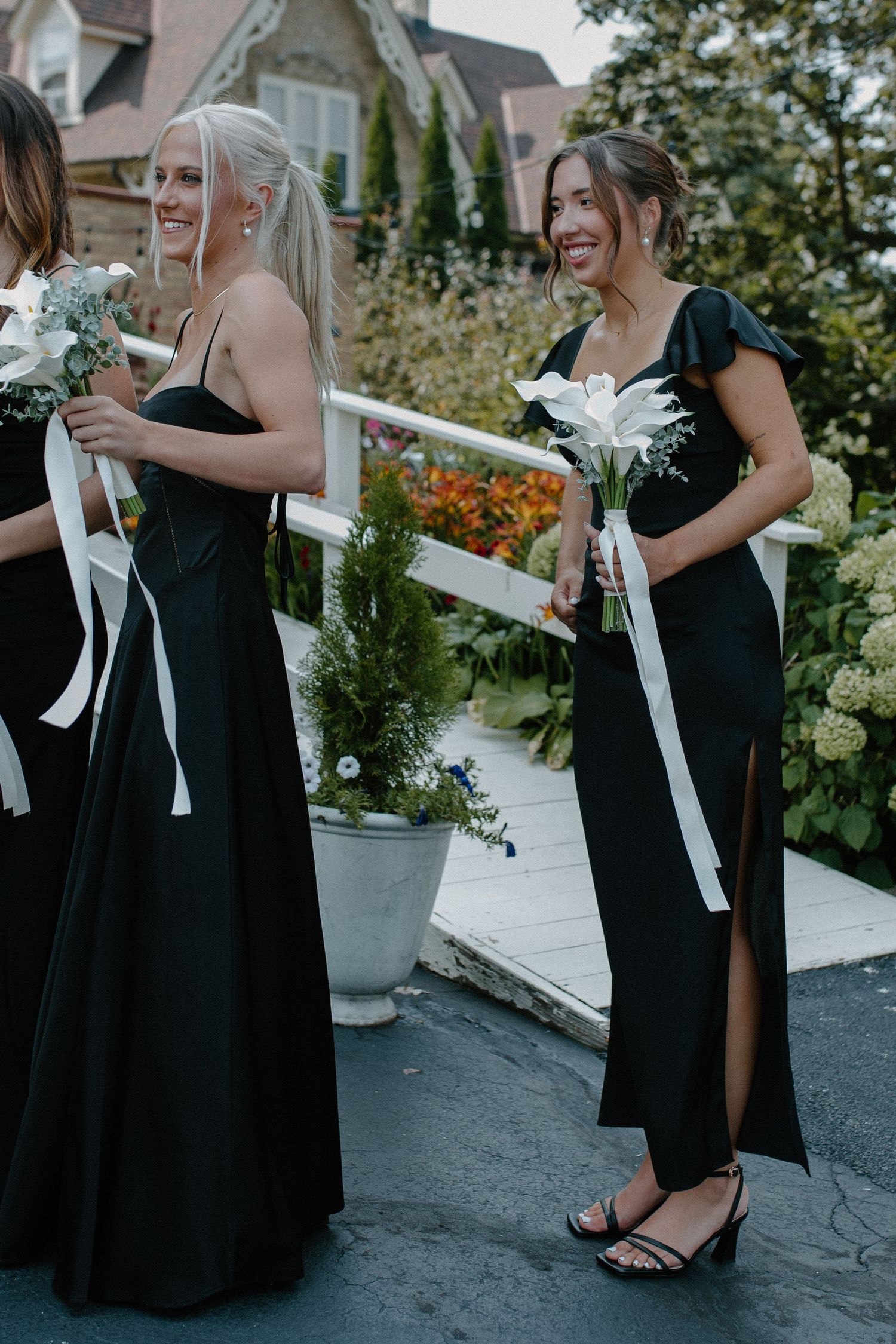 Bridesmaids in elegant black dresses hold white bouquets in a garden setting at Gardens Wedding Center in Wisconsin