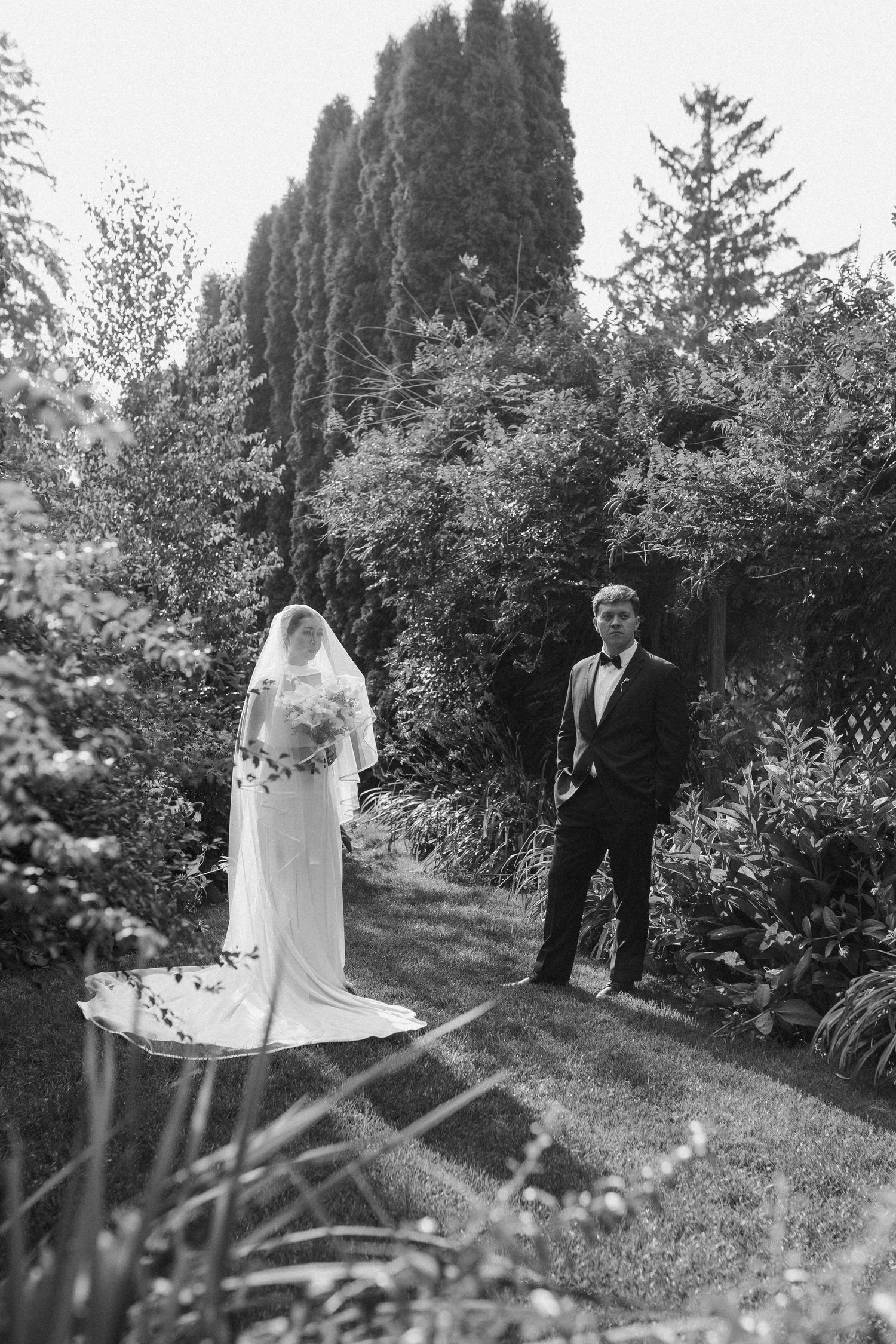 Black and white photo of a bride in a flowing gown and groom in a tuxedo standing in a lush garden.