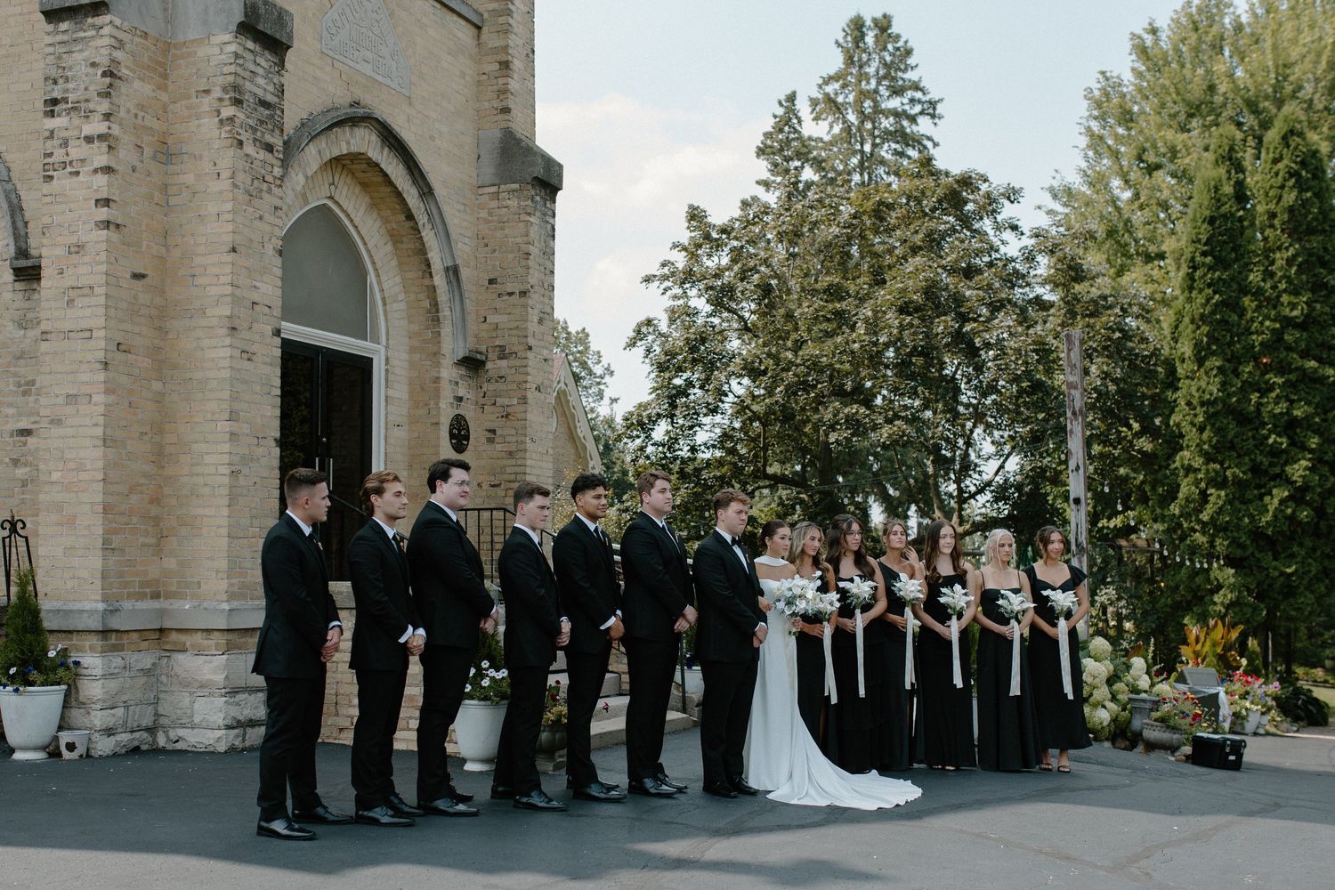 Wedding party lined up in formal black attire outside historic stone church at Gardens Wedding Center in Wisconsin