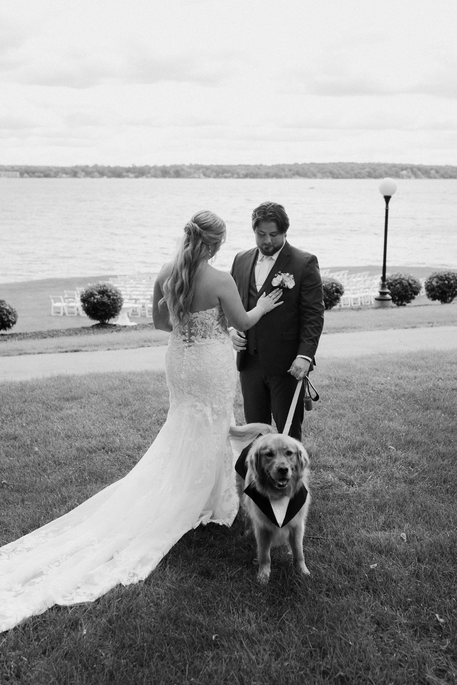 Black and white wedding photo of bride and groom with their dog dressed in a tuxedo by a waterfront.