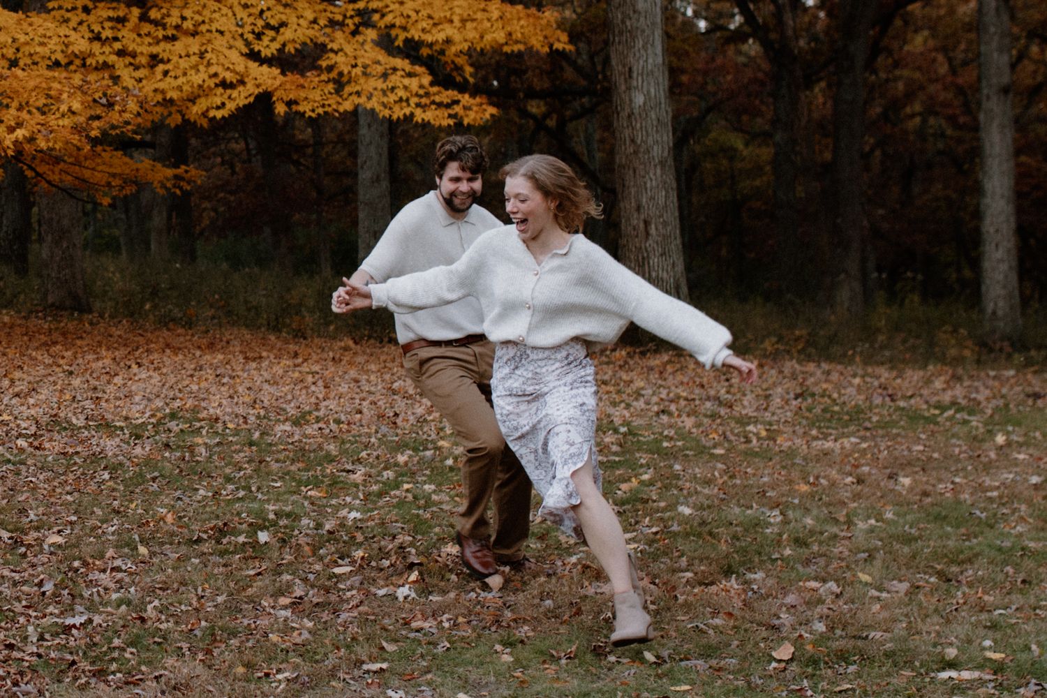 Couple in white and brown outfits chasing each other in a fall forest setting at Lapham Peak engagement setting