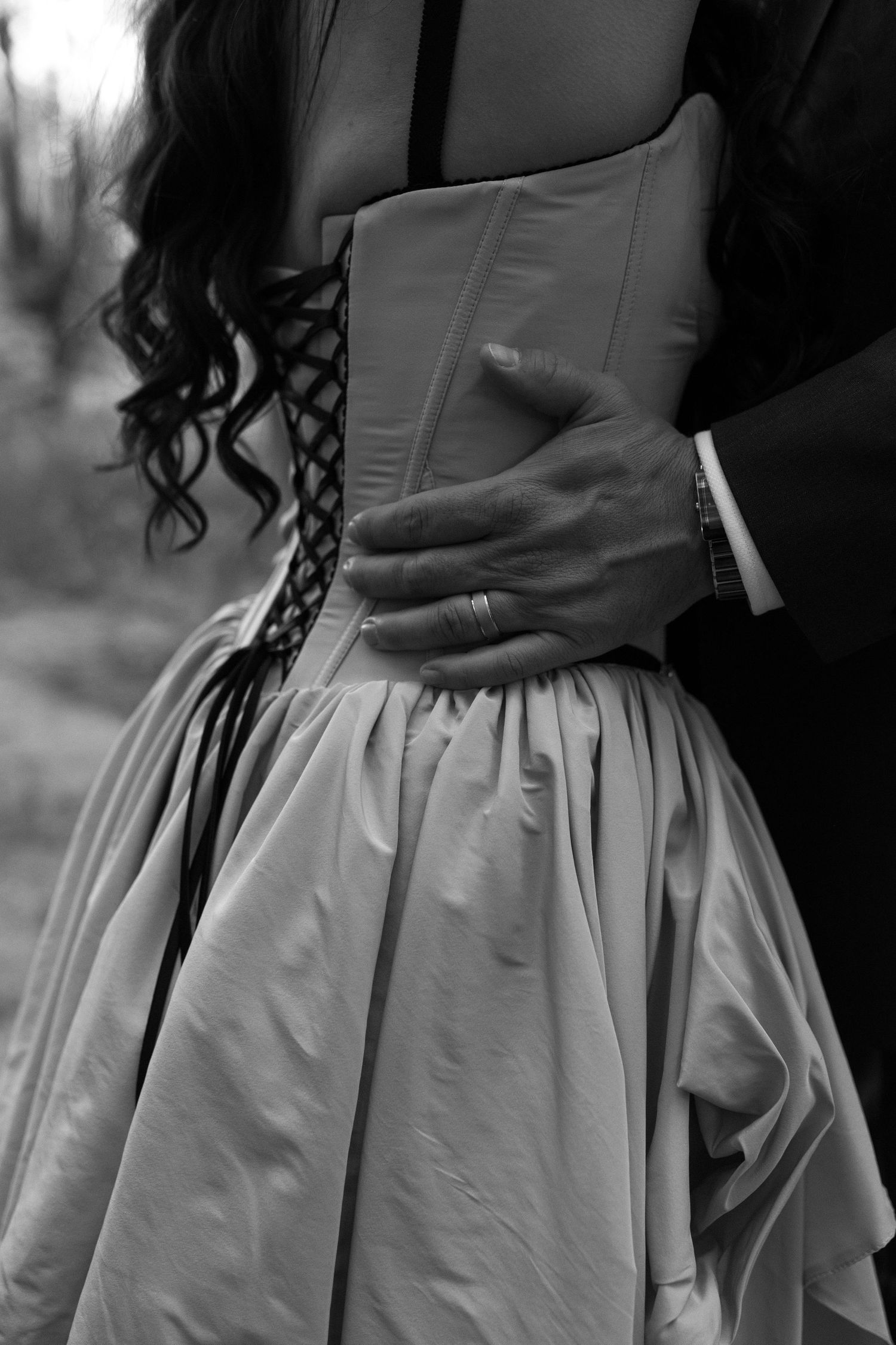 Black and white photo of a couple embracing, mans hand with wedding ring resting on womans corseted wedding dress waist.