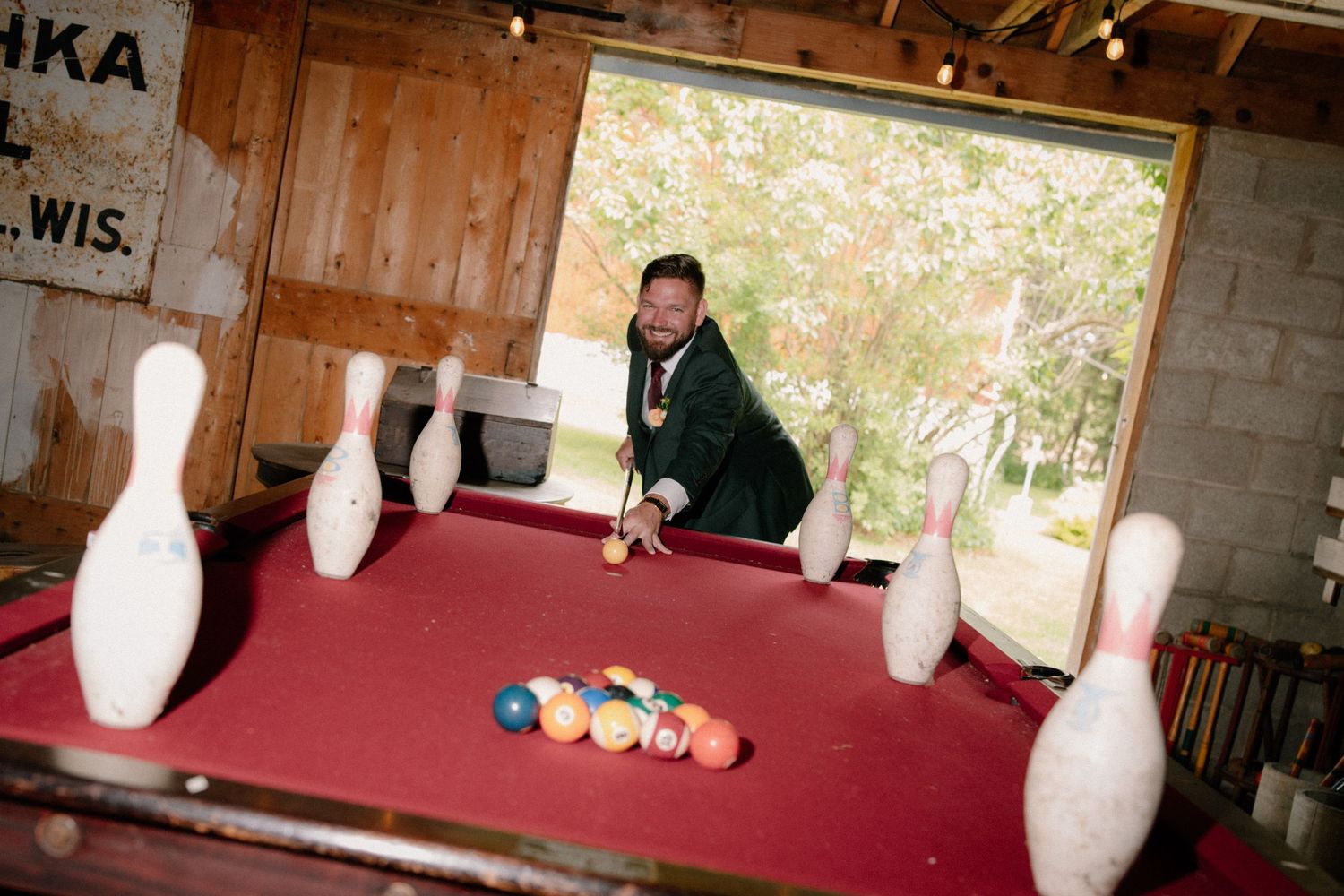 Man smiling while playing pool on a red billiard table surrounded by bowling pins in a rustic game room.