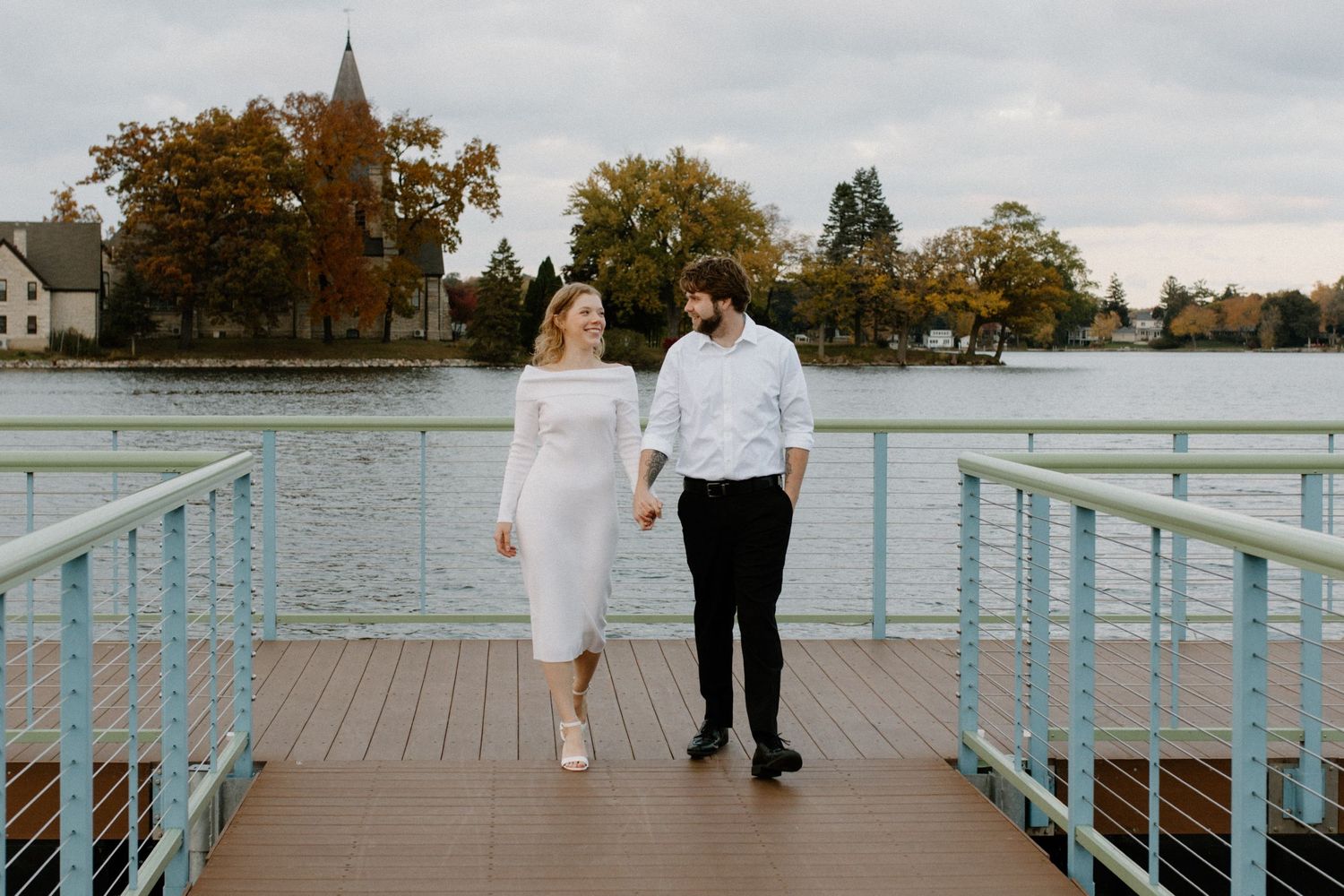 A couple walks along a waterfront boardwalk with railings on an overcast day during Oconomowoc engagement session