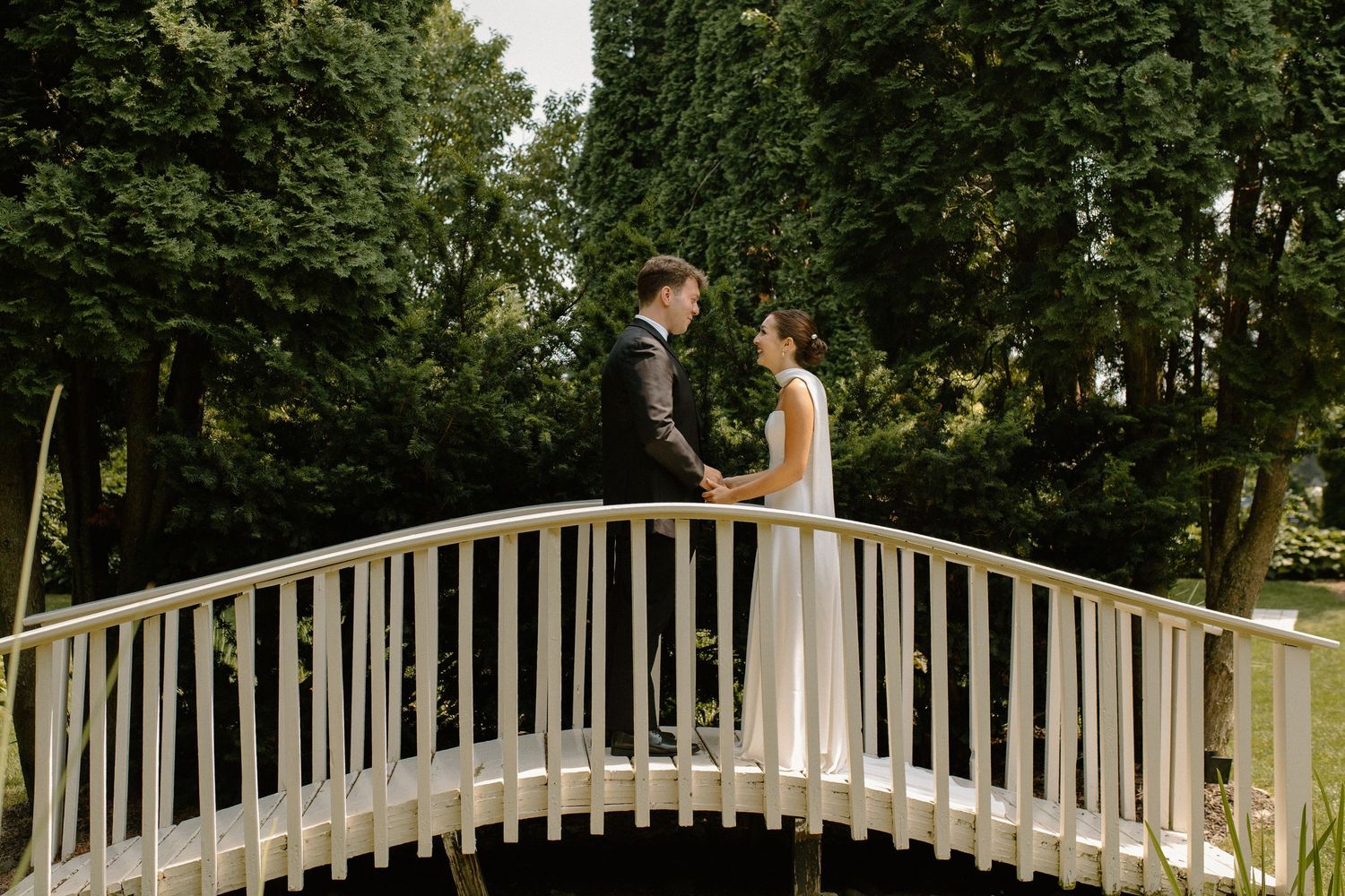 Bride and groom share romantic moment during first look on white wooden bridge surrounded by lush greenery at Gardens Wedding Center in WI