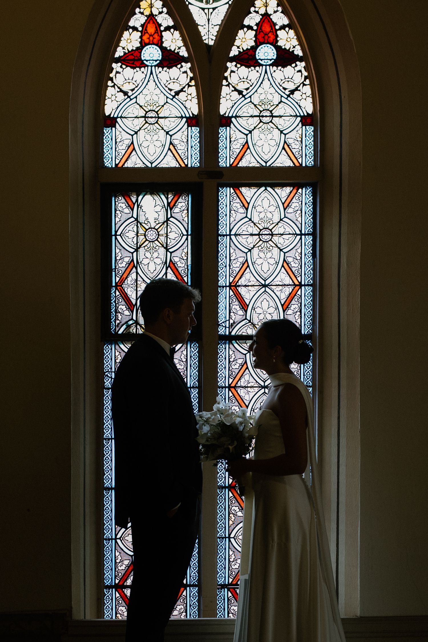 Bride and groom silhouetted against a colorful stained glass church window, bride holding white bouquet.