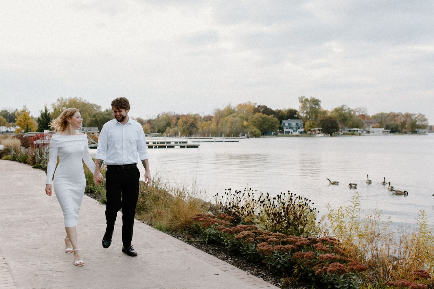 Wide landscape shot of a couple walking along a waterfront path with geese in the distance during Oconomowoc engagement session
