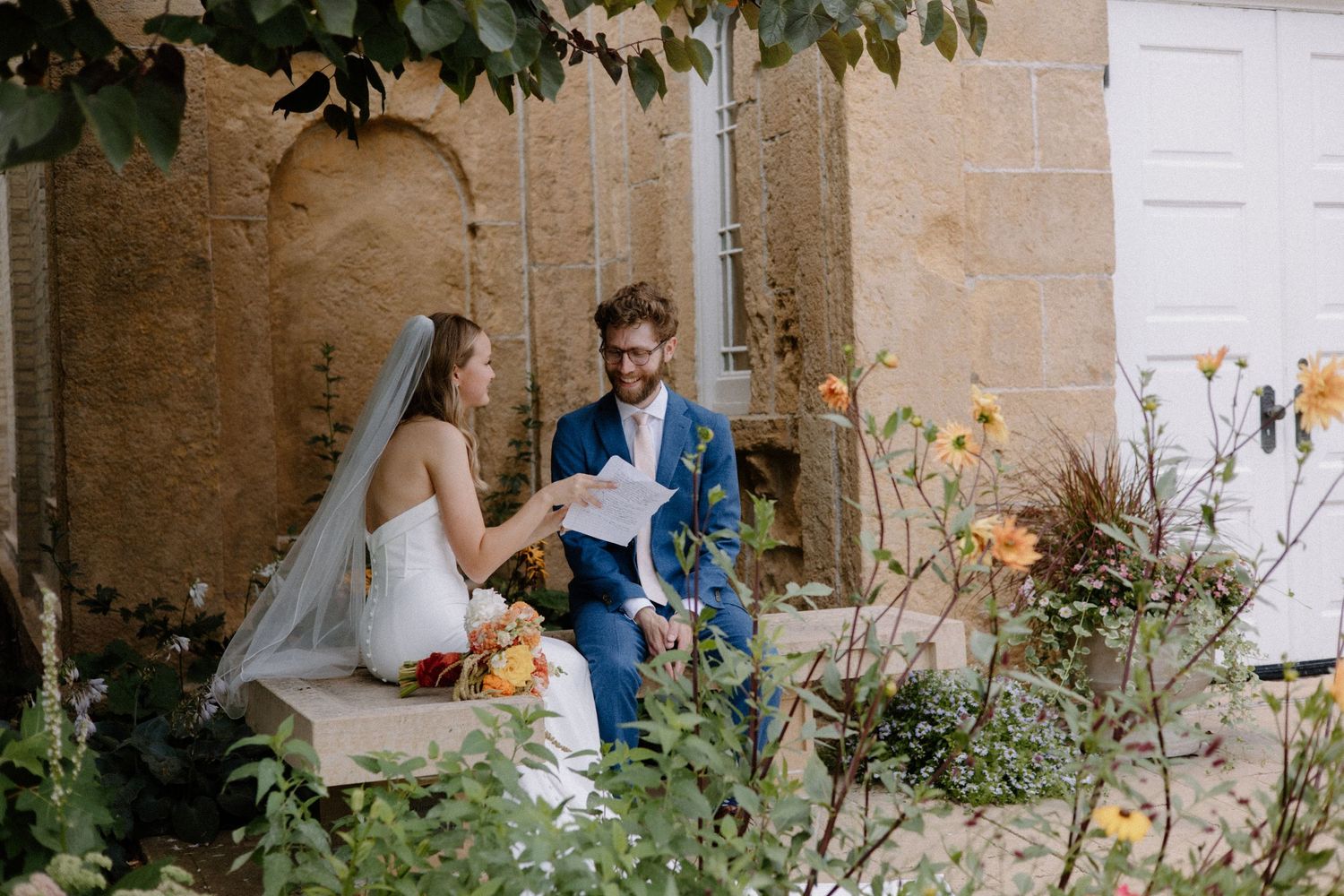 A couple sits in a garden setting during their wedding ceremony surrounded by flowers and greenery in downtown Madison, WI