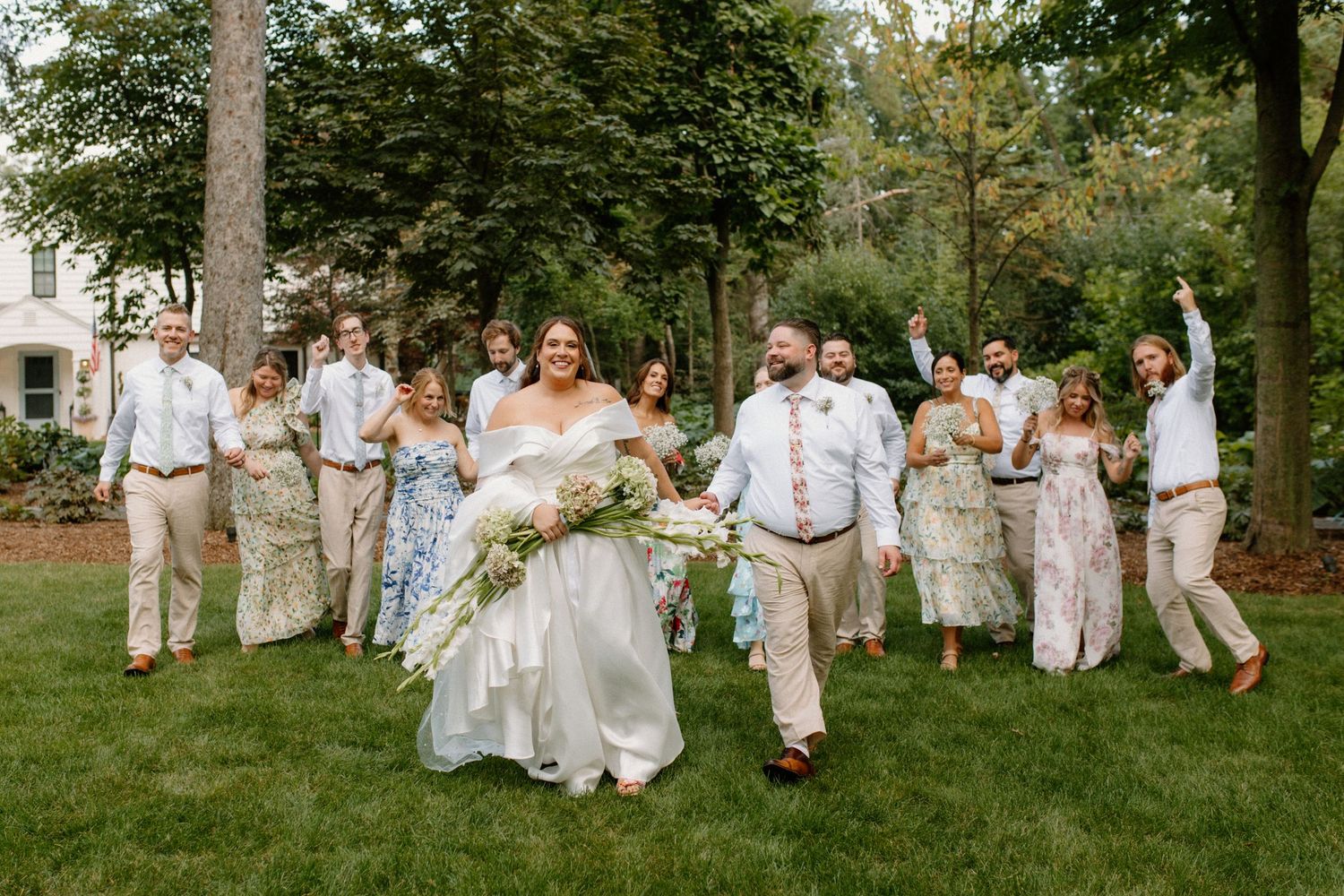 Wedding party celebrating outdoors on green lawn, bride in white gown holding bouquet, groom and guests in festive attire.