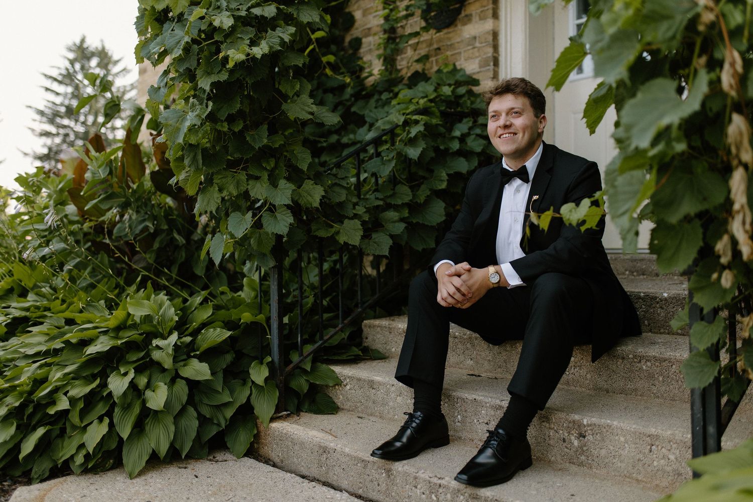 Groom in black tuxedo sitting relaxed on stone steps surrounded by lush green ivy at outdoor wedding venue.