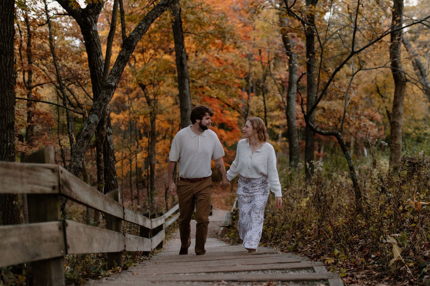 A couple walks hand in hand along a wooden walkway through autumn trees at Lapham Peak engagement session