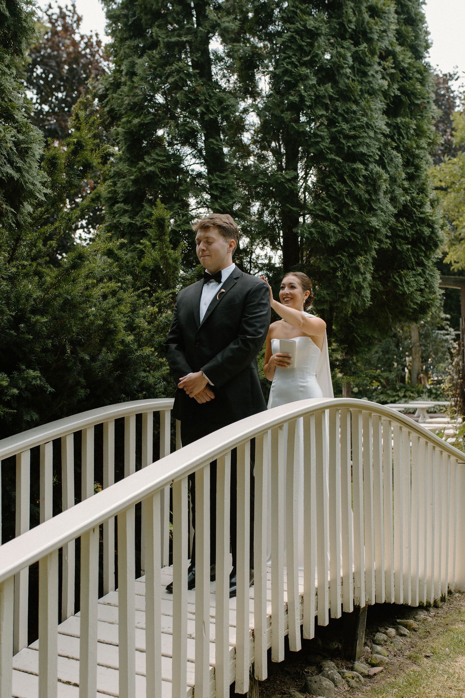 A couple stands on a white arched bridge surrounded by tall evergreen trees during their garden wedding day in Wisconsin