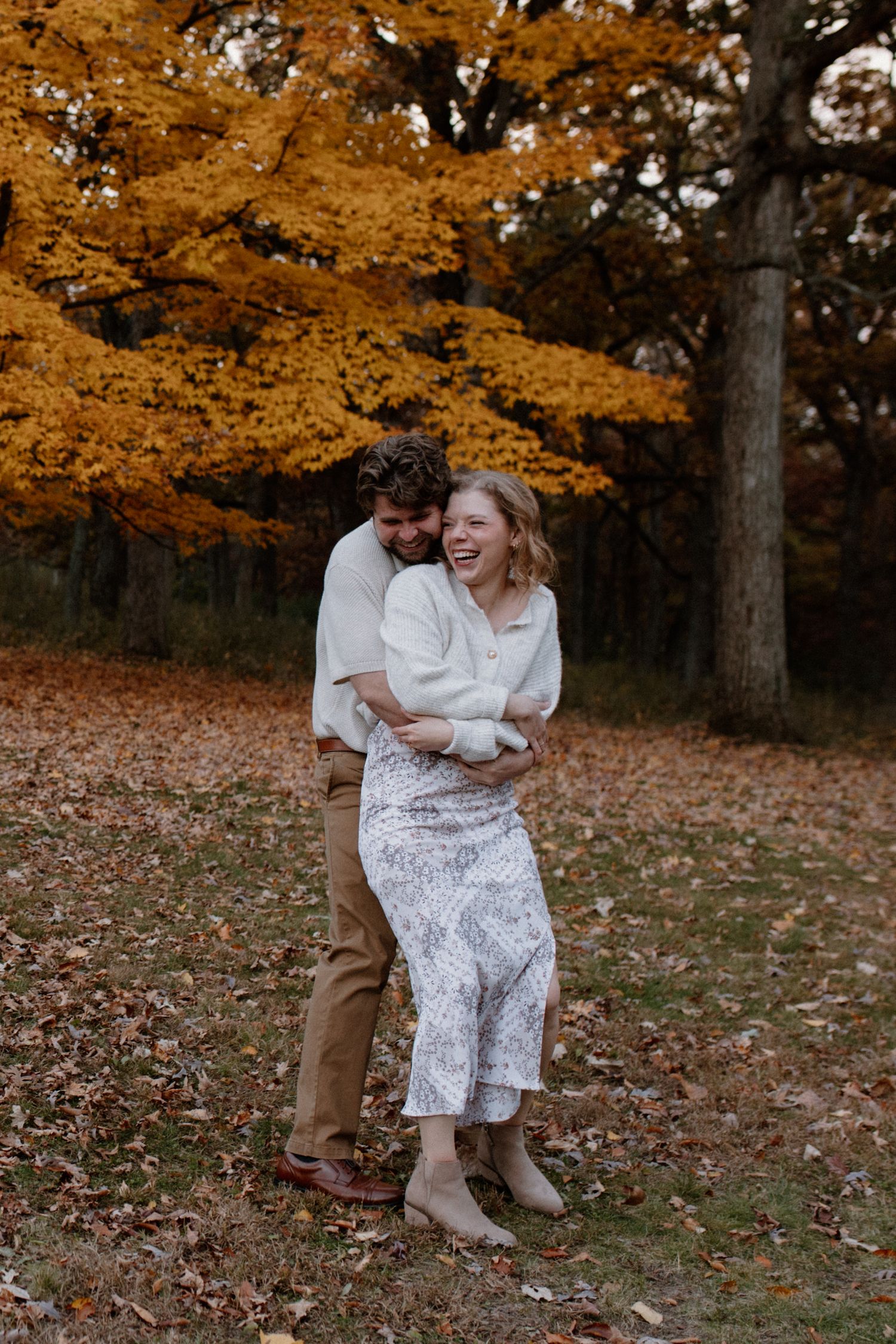 A couple embraces under vibrant orange autumn trees with fallen leaves scattered around at Lapham Peak engagement session