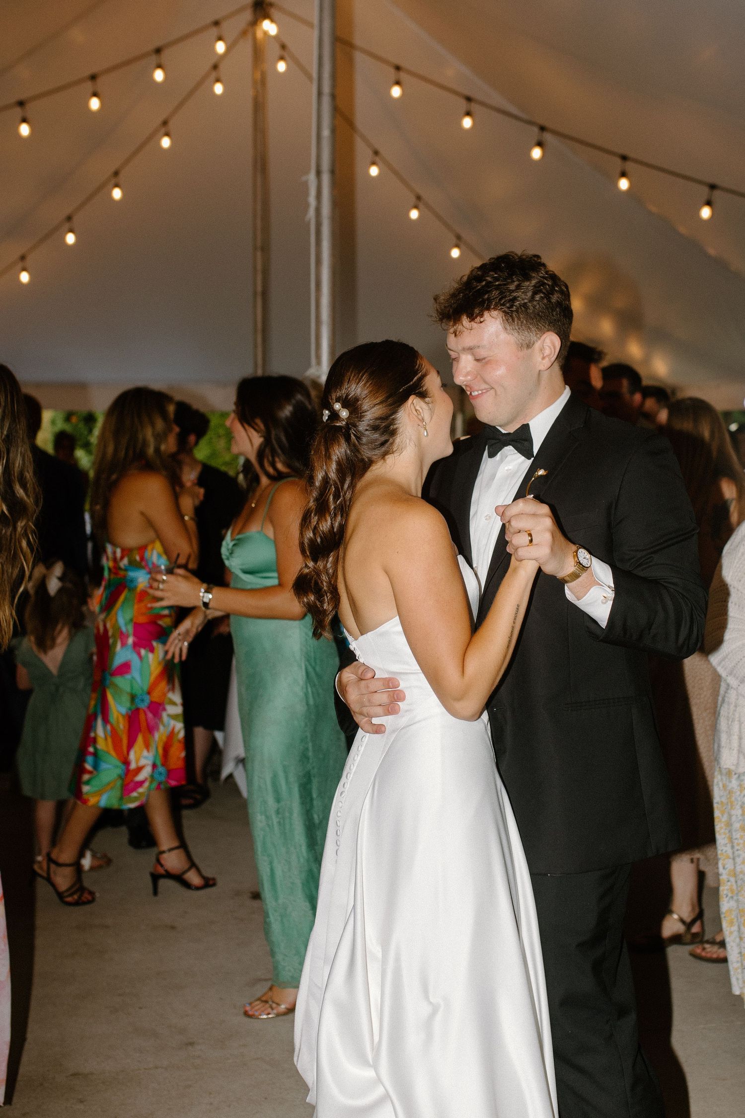 Bride and groom share their first dance under string lights in a white tent at their elegant wedding reception.