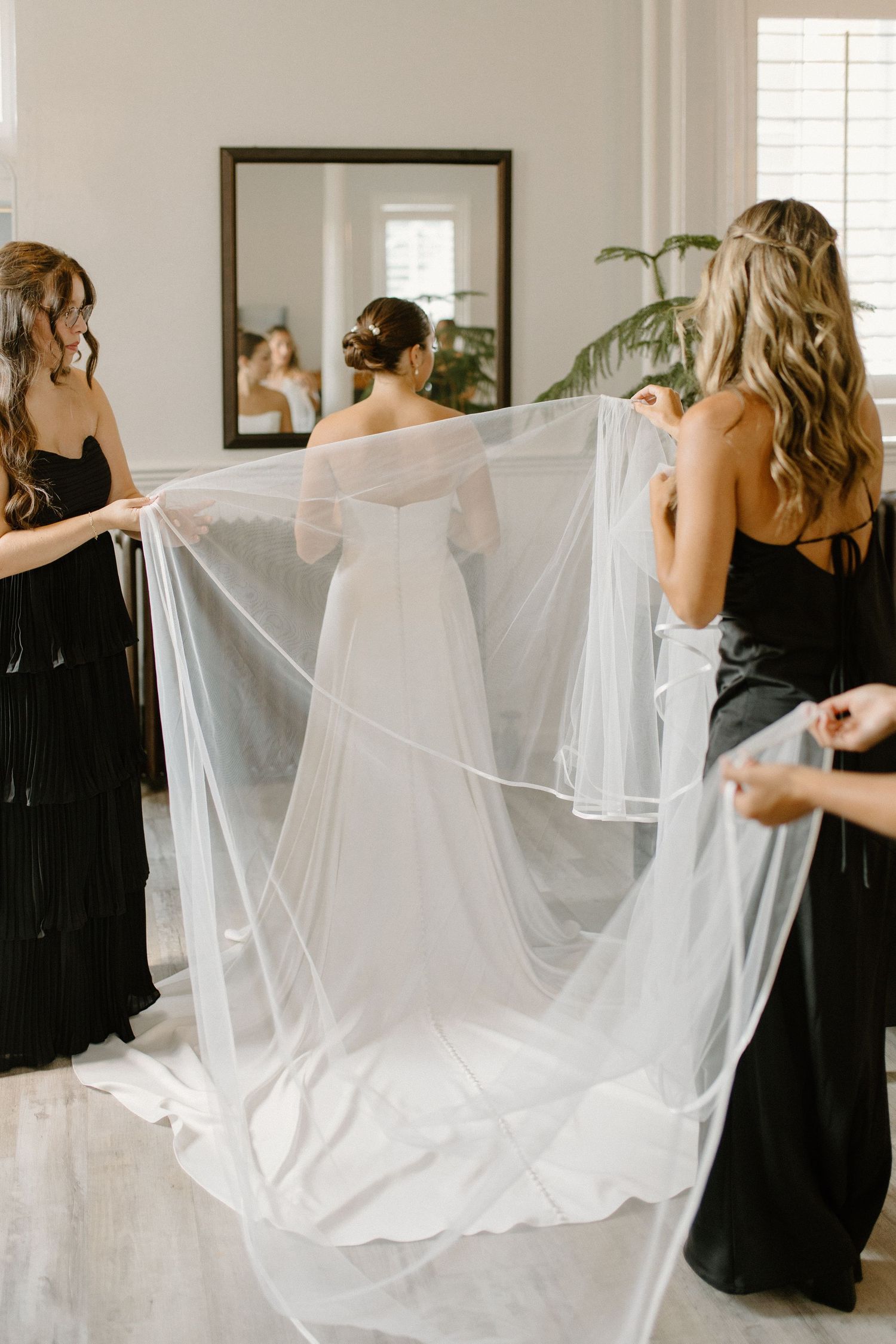 Bridesmaids in black dresses hold a bride's long white veil as she stands in her wedding gown before a mirror.