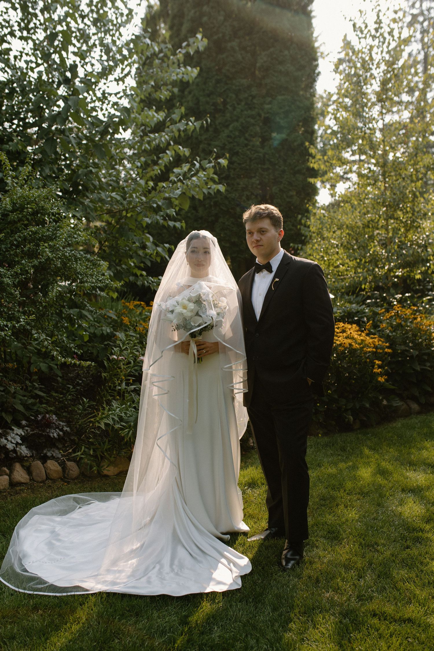 Bride in white gown and veil holding bouquet stands with groom in black tuxedo in lush garden setting.