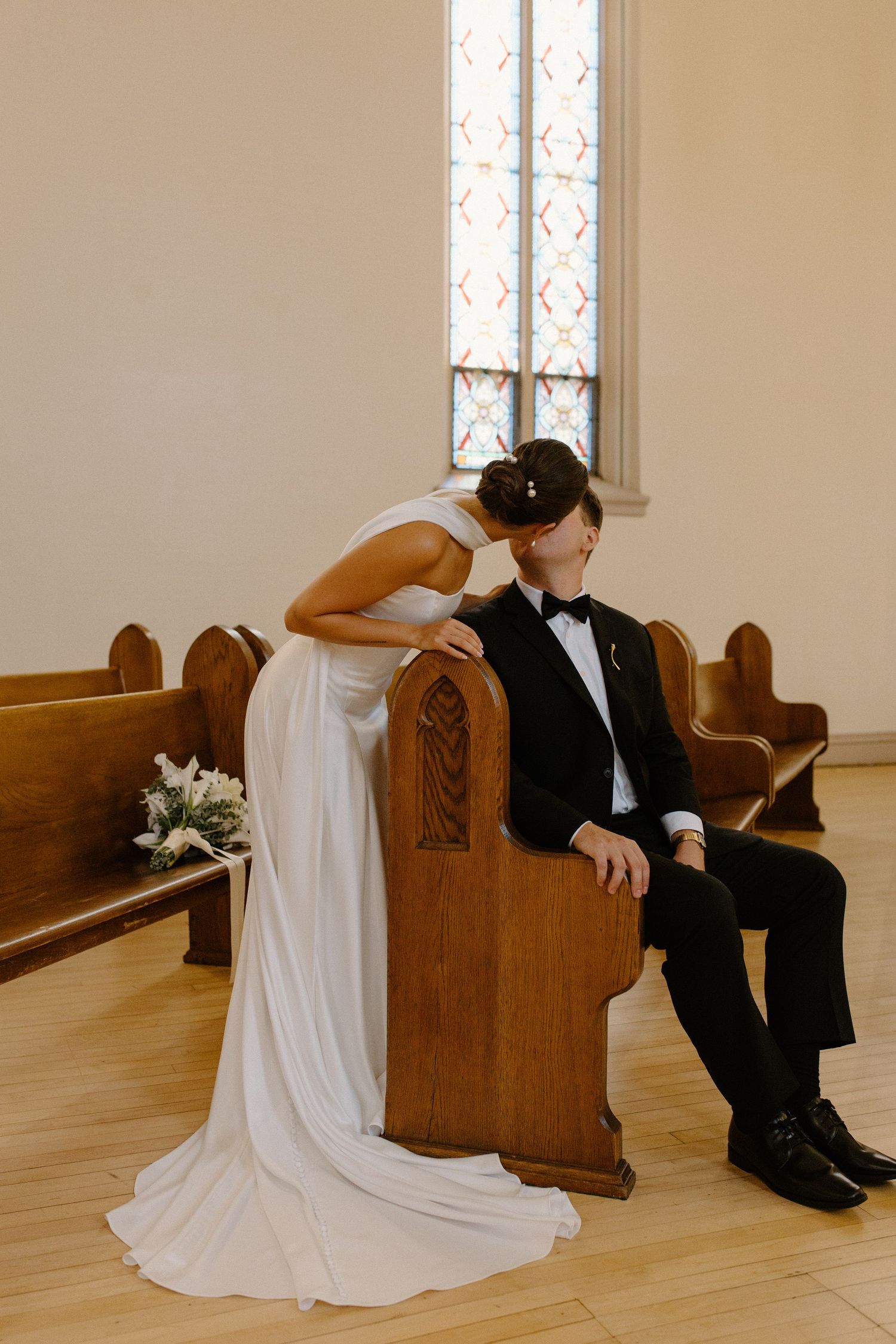 Bride in white gown kisses groom in black tuxedo seated in wooden church pew during wedding ceremony.