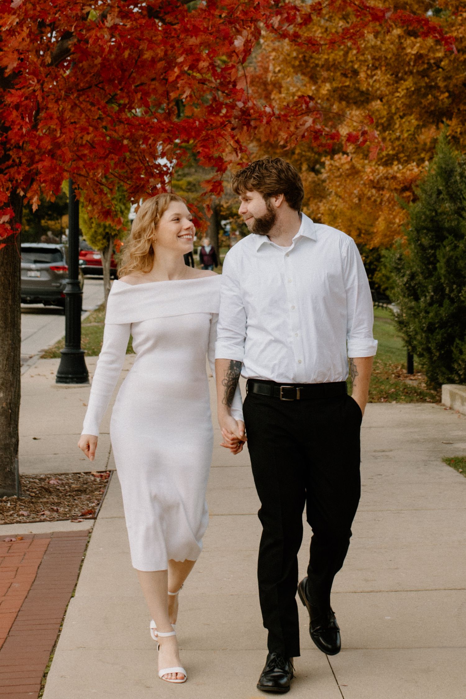 A couple walks hand in hand under beautiful autumn trees with red leaves in downtown Oconomowoc engagement session