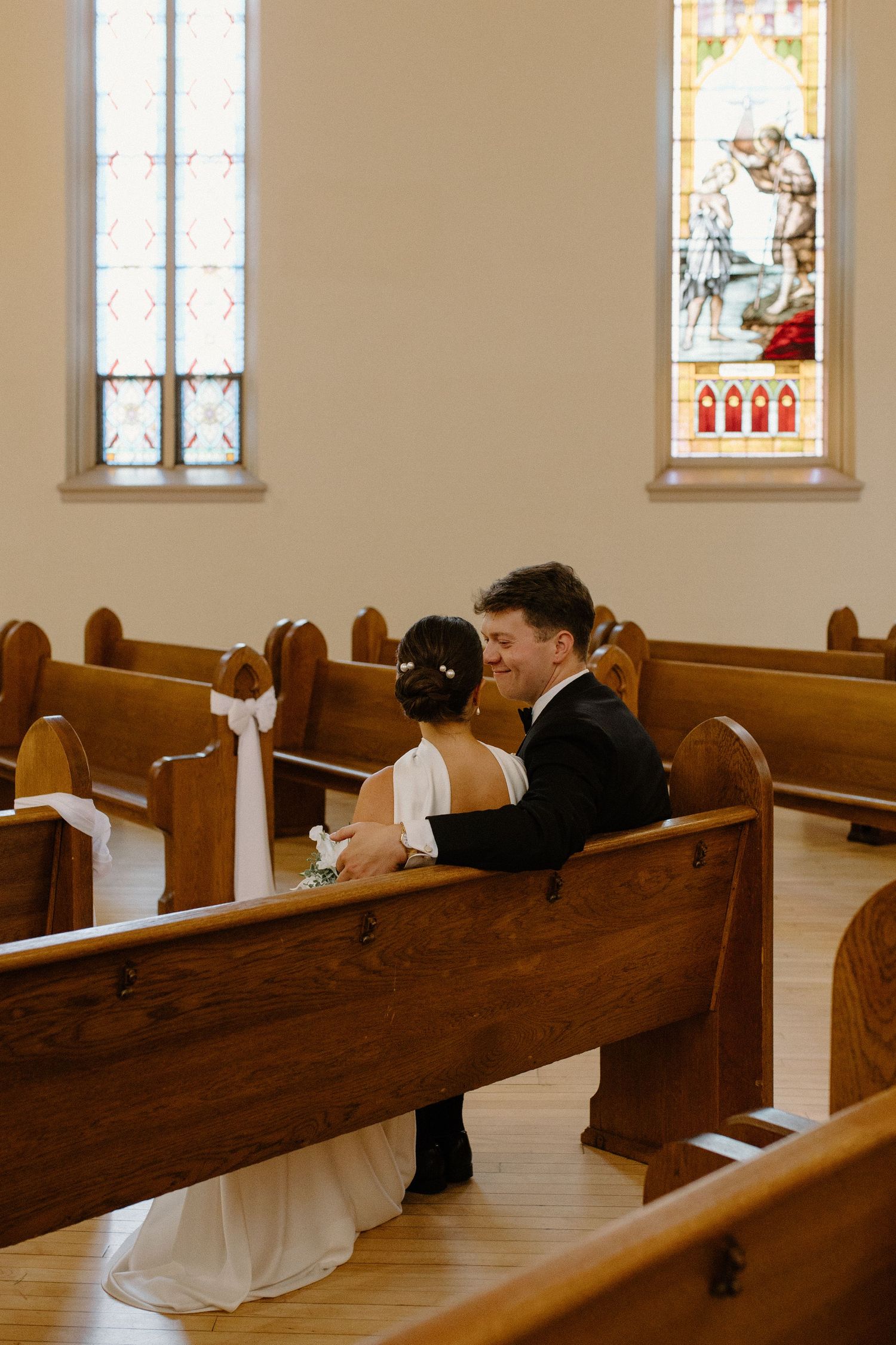 A bride and groom share a joyful moment seated together in a sunlit church pew with stained glass windows.