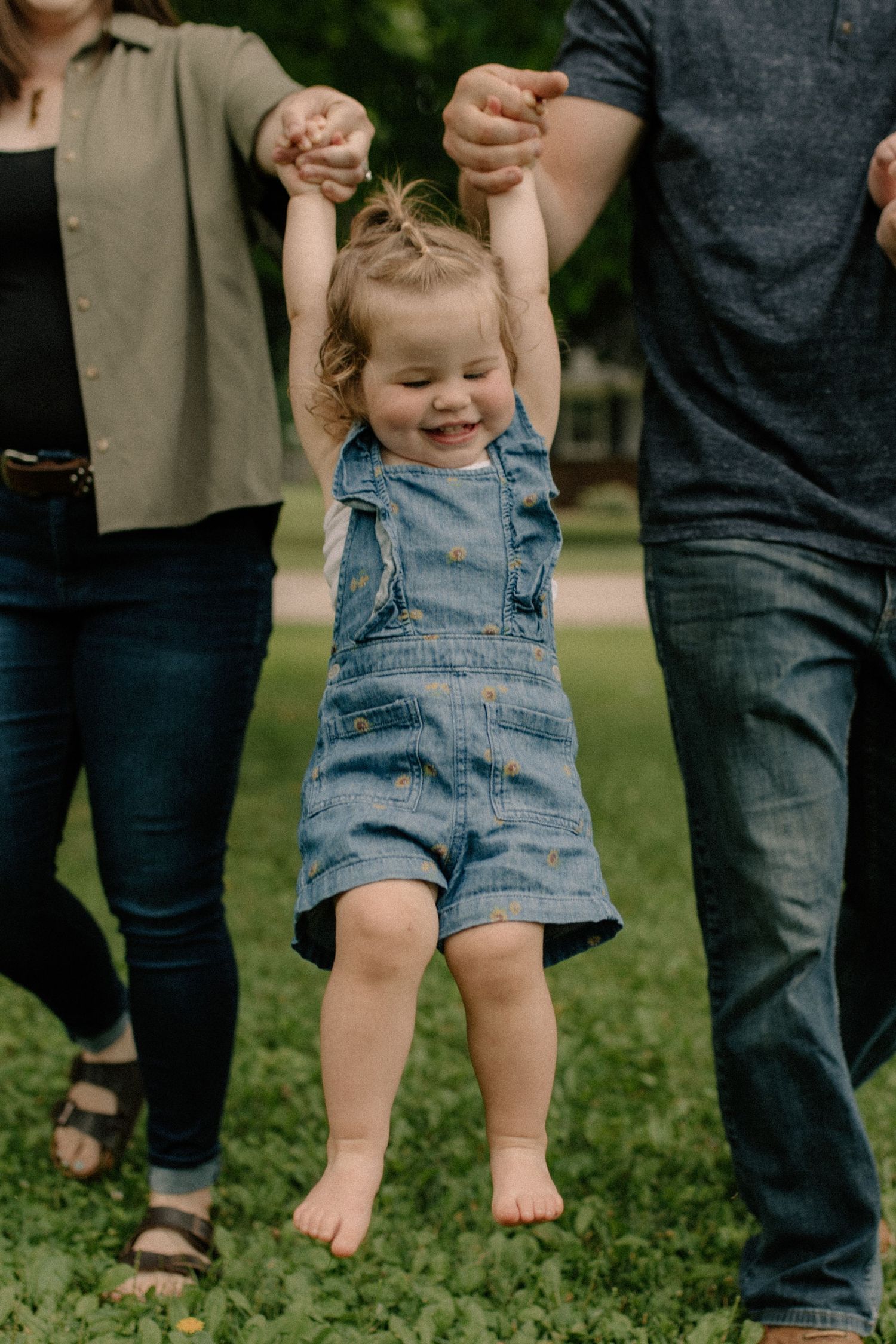Small child in denim overalls being swung between two adults for candid family photos