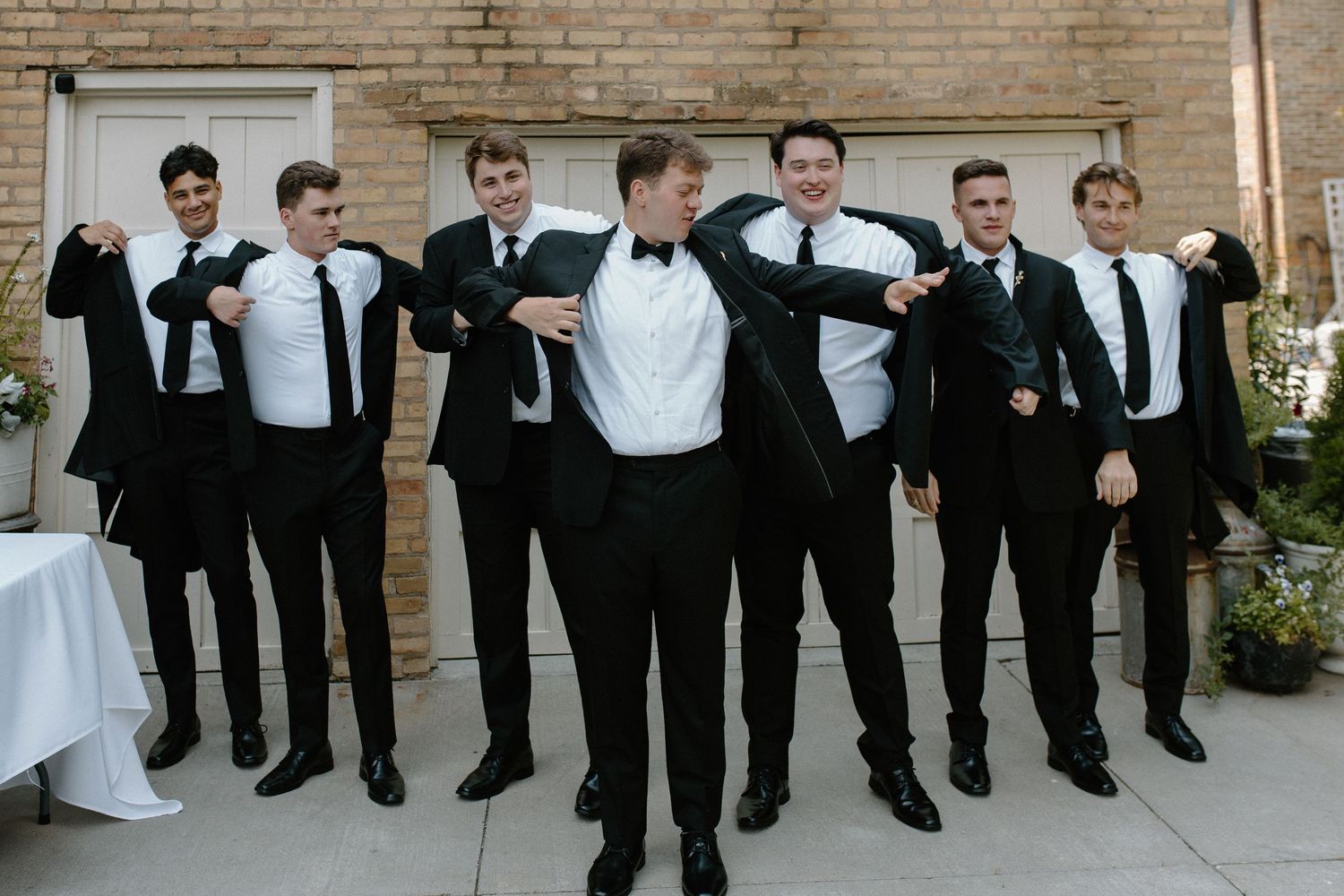 Groomsmen lined up against brick wall wearing black tuxedos at garden wedding in Wisconsin