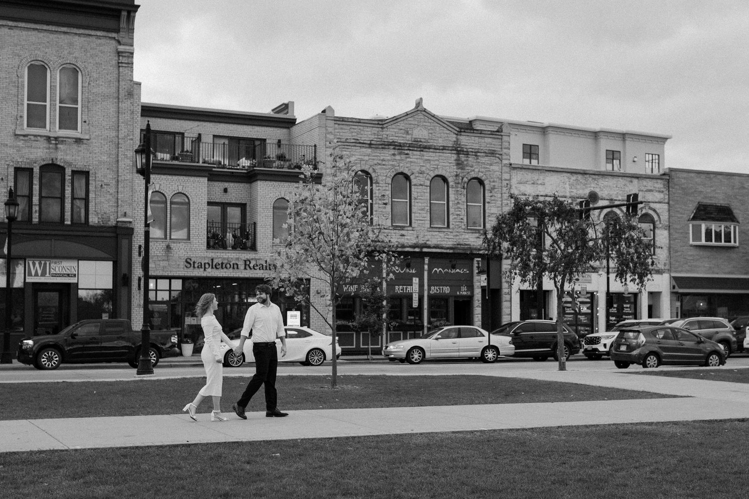 A couple walks together in black and white panoramic view of downtown Oconomowoc during engagement session