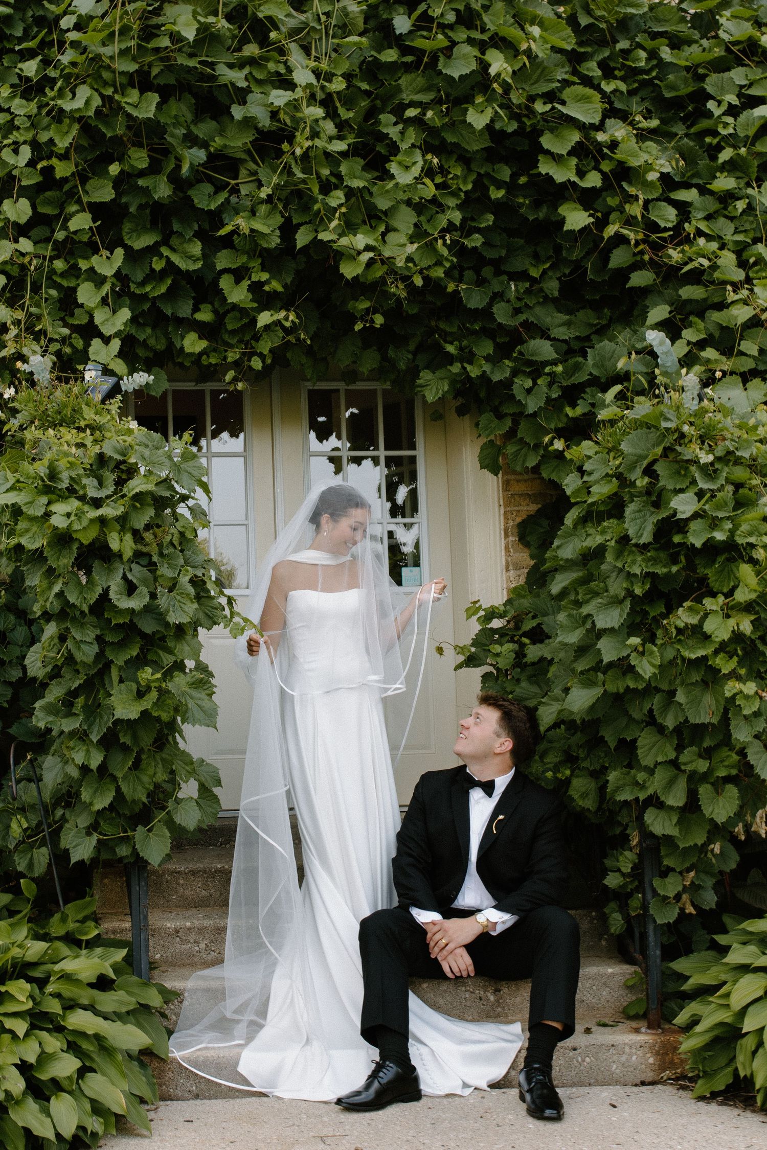 Bride in white gown and veil stands beside groom in tuxedo seated on steps, framed by lush green ivy-covered wall.