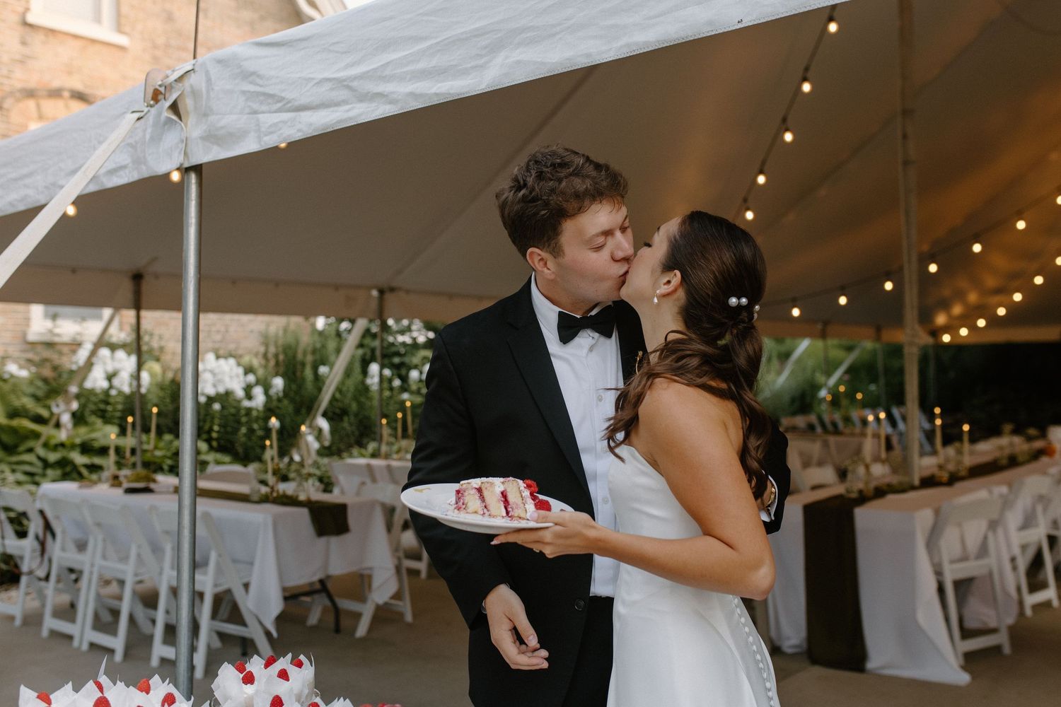 Newlywed couple sharing a kiss under a white tent at their elegant outdoor wedding reception while holding a cake.