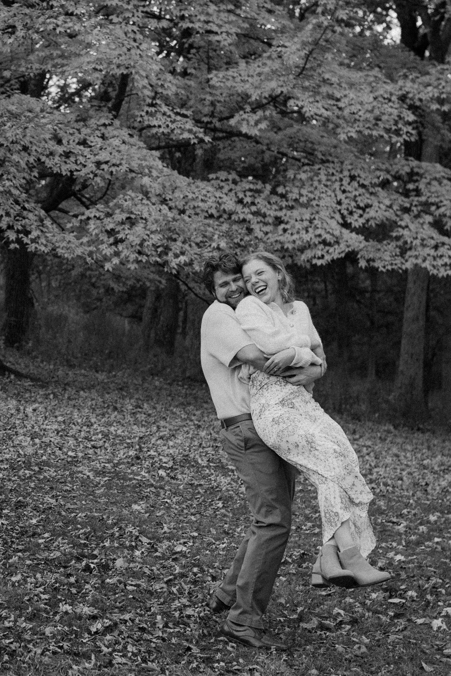 A playful black and white photo of a couple embracing in a leaf-covered forest at Wisconsin engagement session