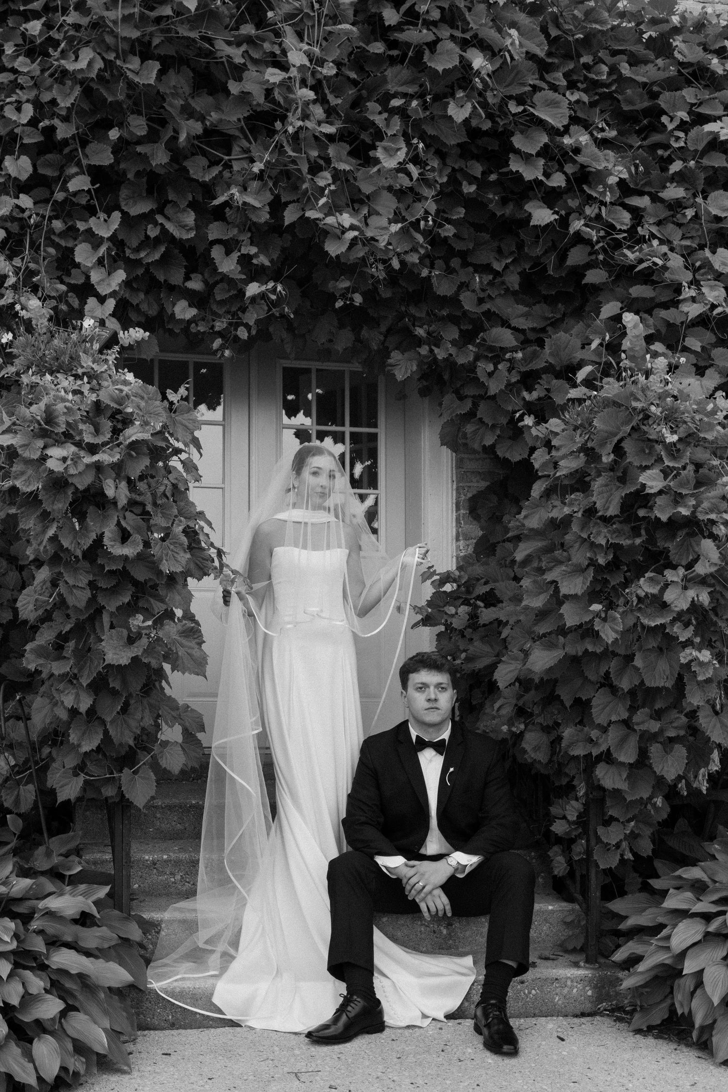 Black and white wedding photo of bride in veil and groom in tuxedo posing by ivy-covered doorway.