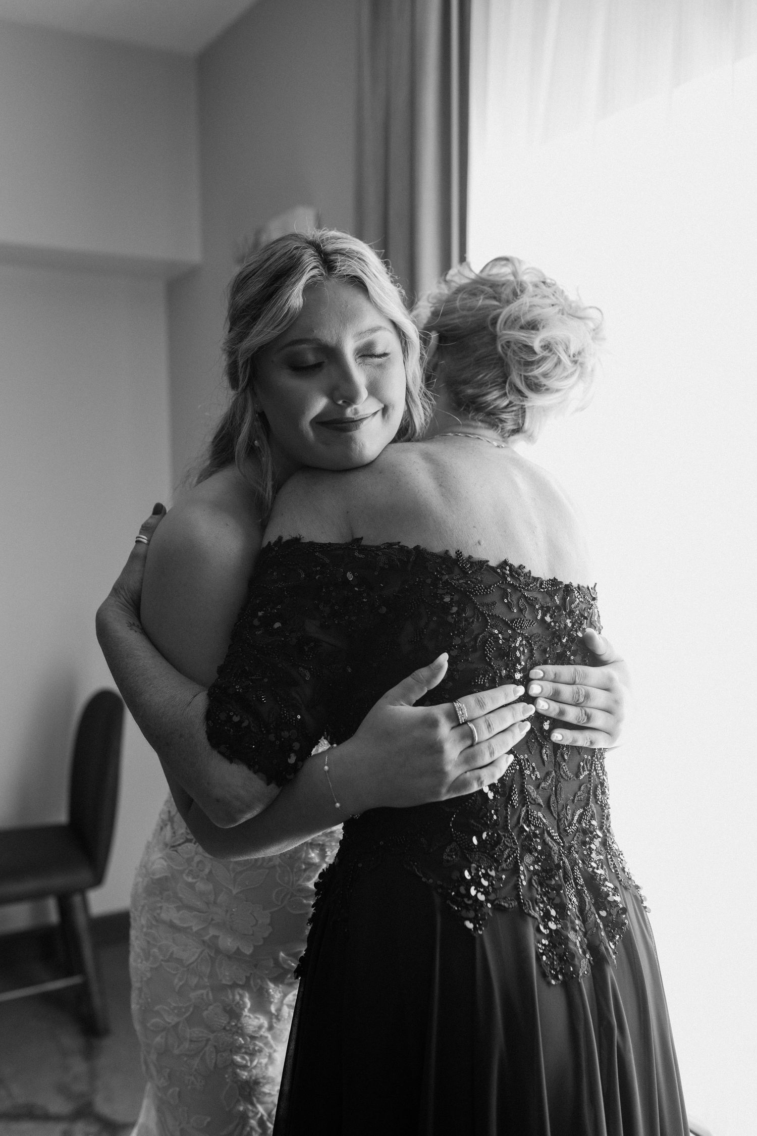 Bride in lace gown shares emotional hug with woman in beaded off-shoulder dress near window, black and white wedding photo.