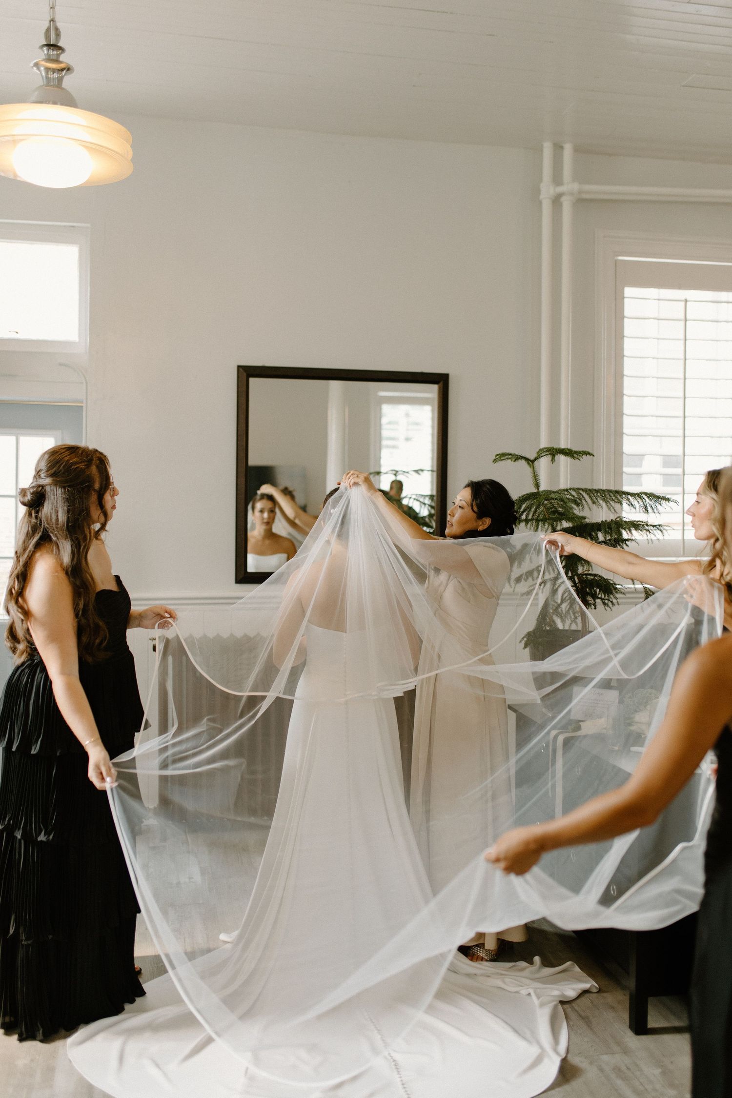 Bridesmaids in black dresses help a bride with her flowing white veil in a bright, elegant bridal suite.