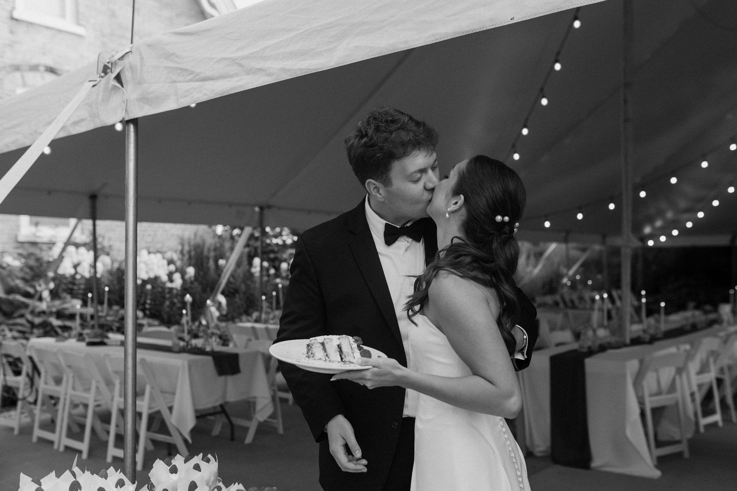 Black and white photo of a bride and groom sharing a kiss while holding wedding cake under a tent.