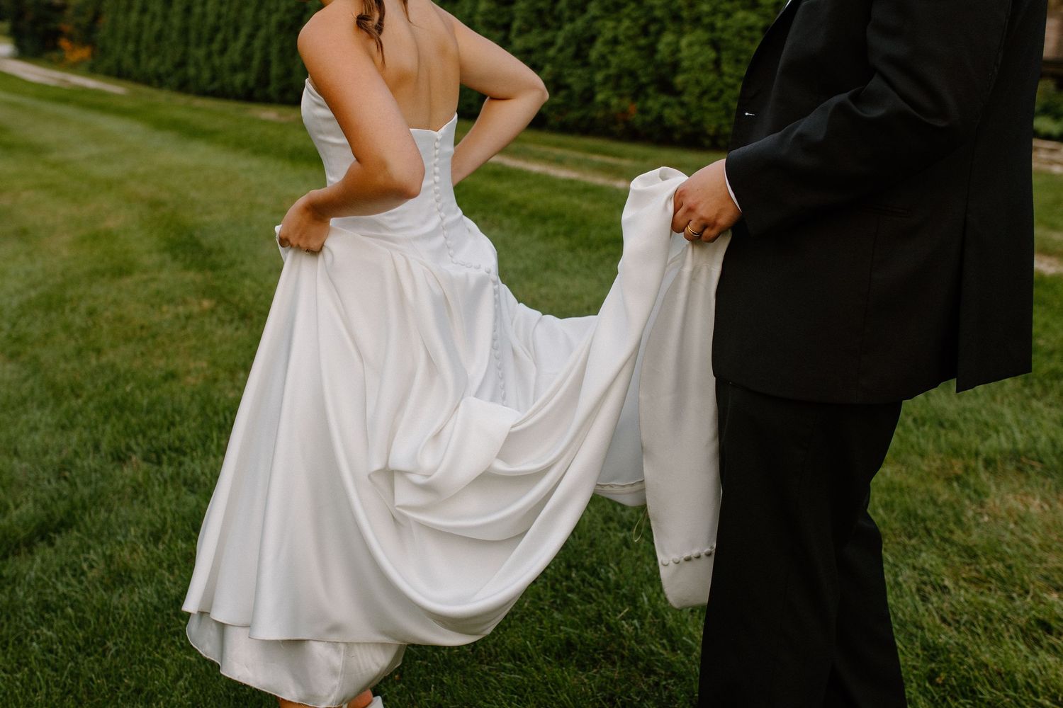 Groom in black suit holding bride's white wedding gown train on green grass at outdoor wedding ceremony.
