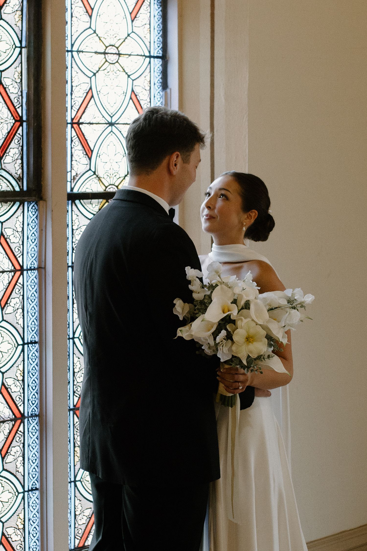 Bride and groom share a tender moment by stained glass windows, bride holding white floral bouquet in elegant wedding attire.