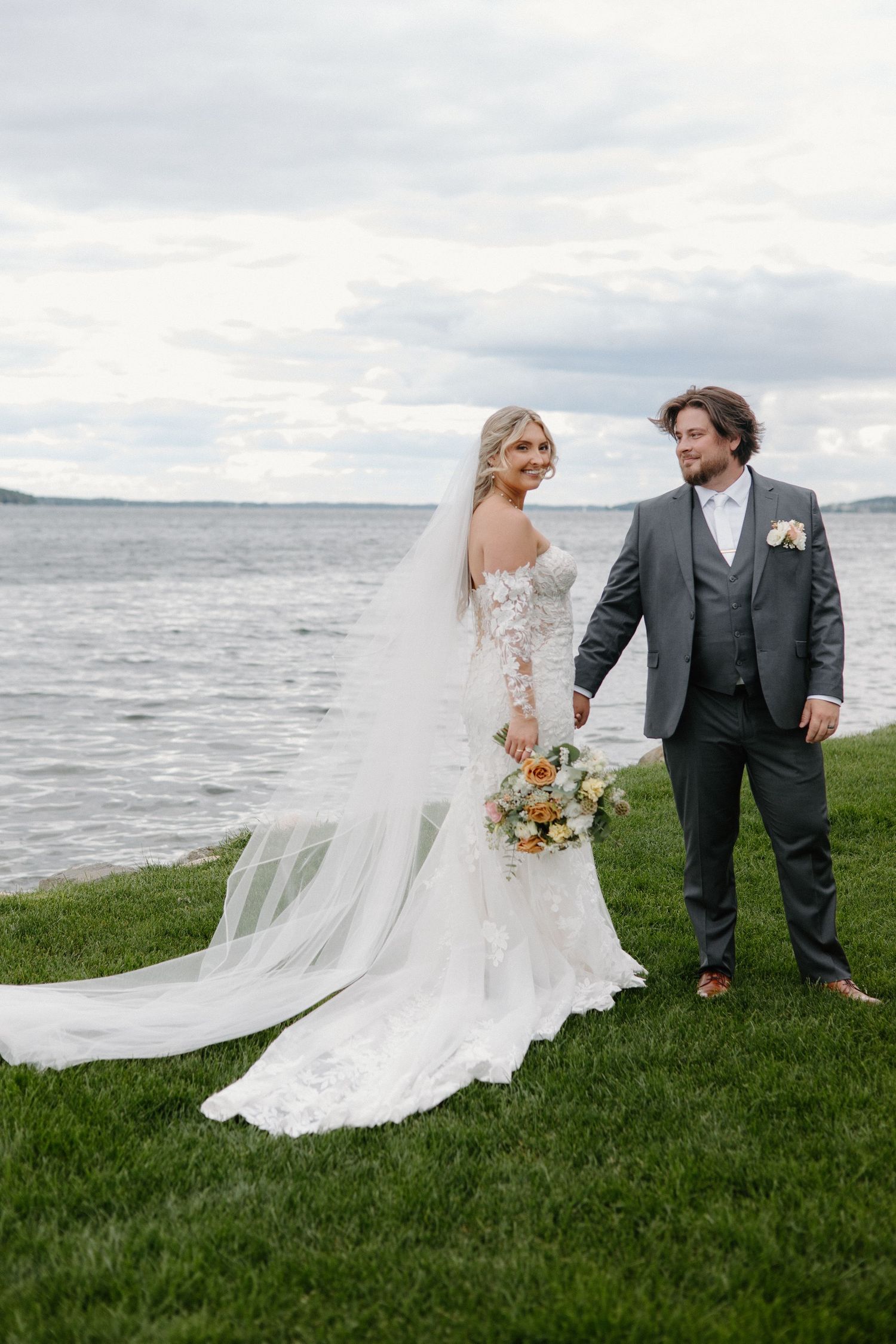Bride in lace gown with flowing veil holds bouquet beside groom in gray suit, posing by waterfront on green lawn.
