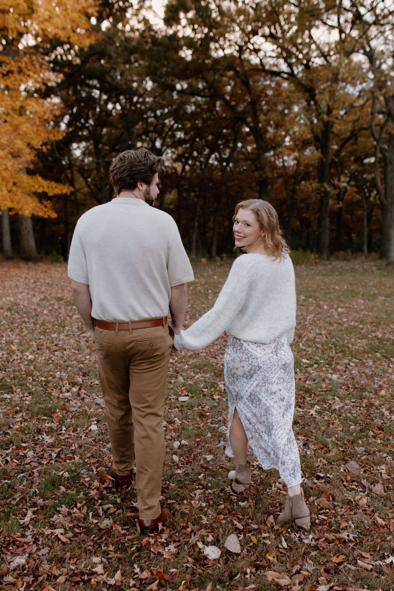 A couple holds hands while walking through fallen autumn leaves in a forest setting during Wisconsin engagement session
