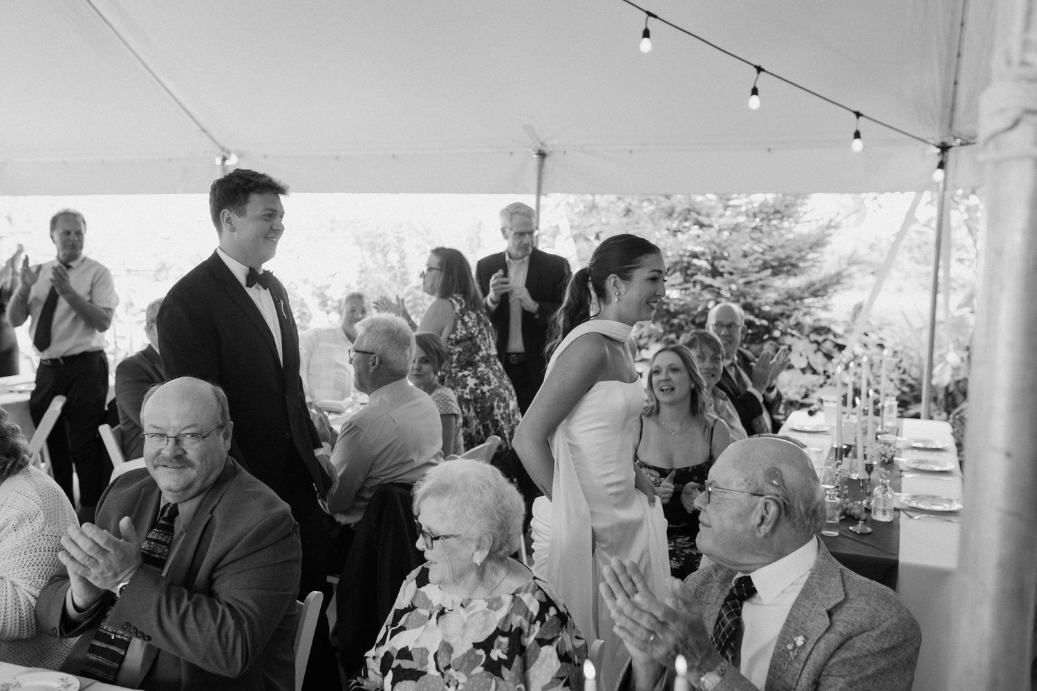 Black and white wedding reception photo showing guests clapping under a tent as the bride and groom share a moment.