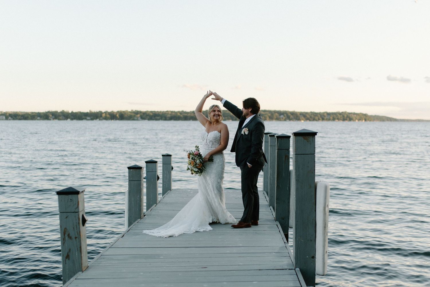 Bride and groom dancing on a wooden dock over a serene lake at sunset during their waterfront wedding.